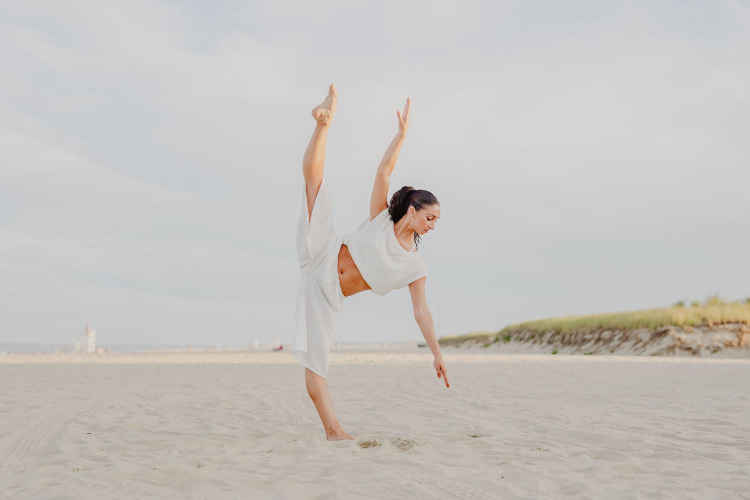 A graceful dancer performs a high leg extension on a sandy beach wearing a flowing white outfit under a cloudy sky.