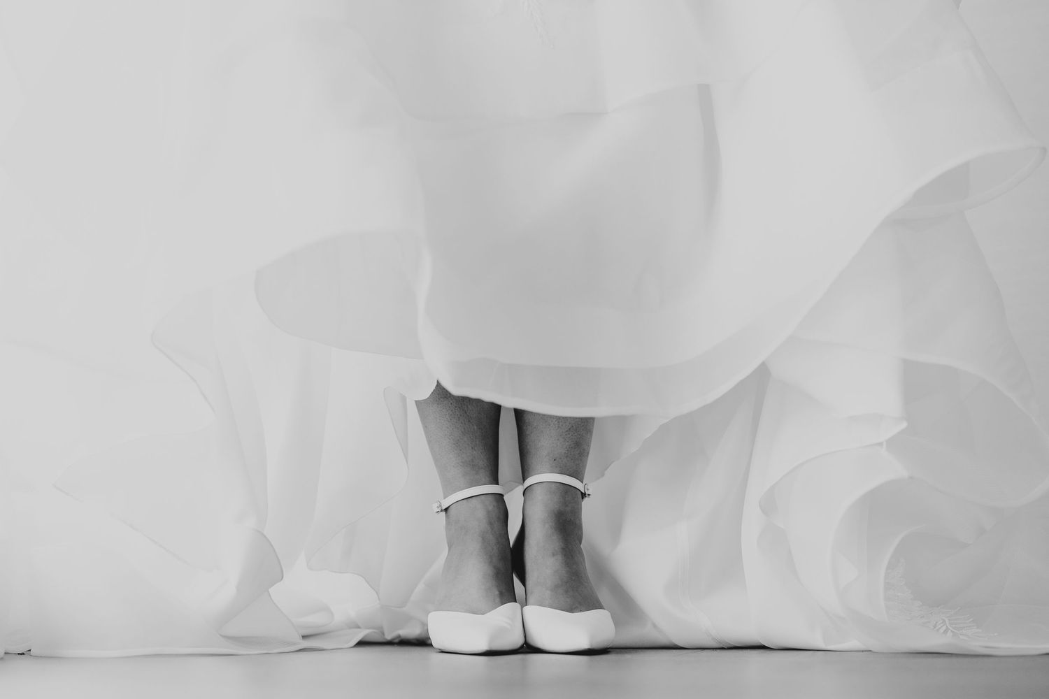 Close-up view of white wedding dress hem and bridal shoes on light background.