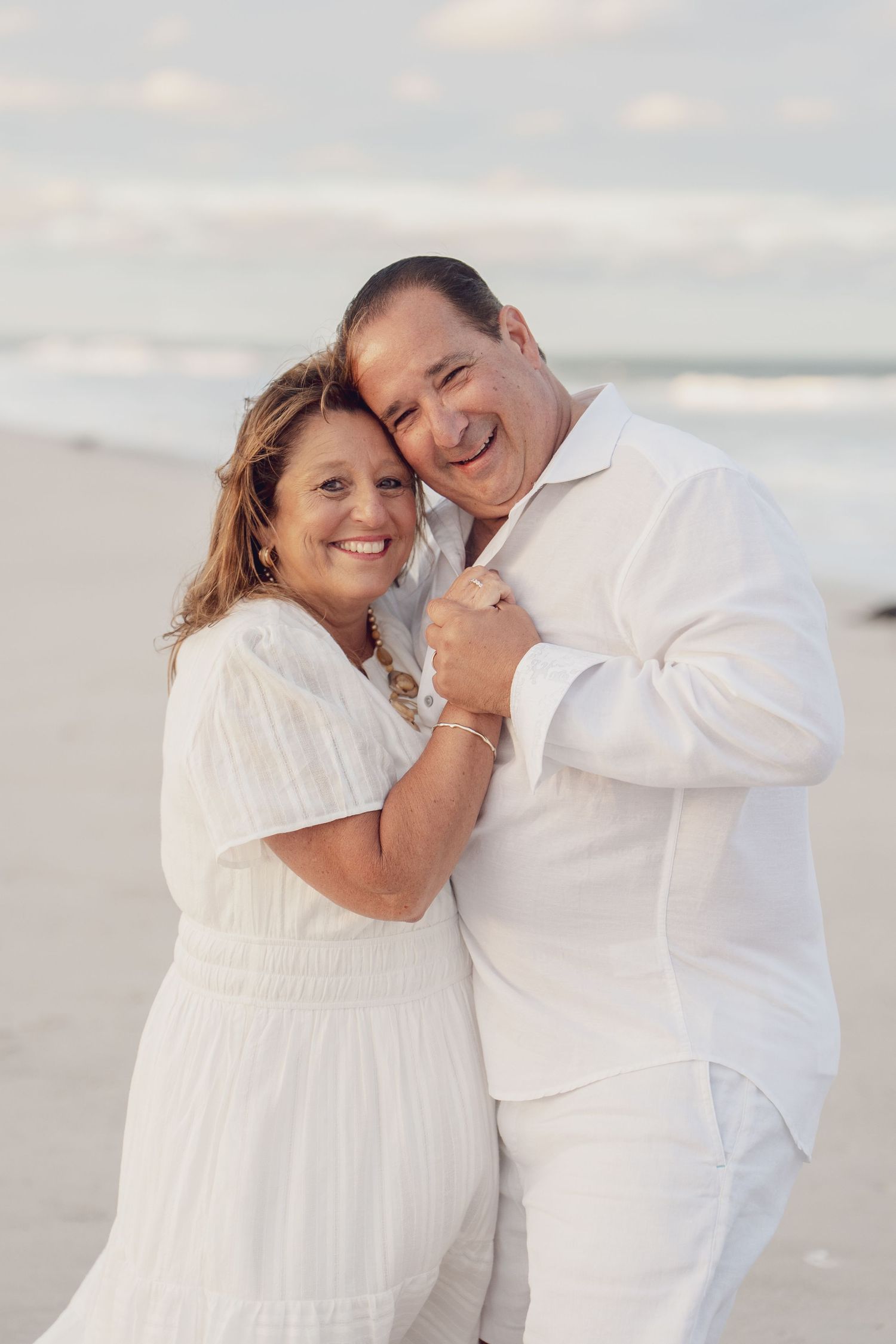 A couple shares an intimate embrace while wearing white outfits on a beach during a romantic photoshoot.