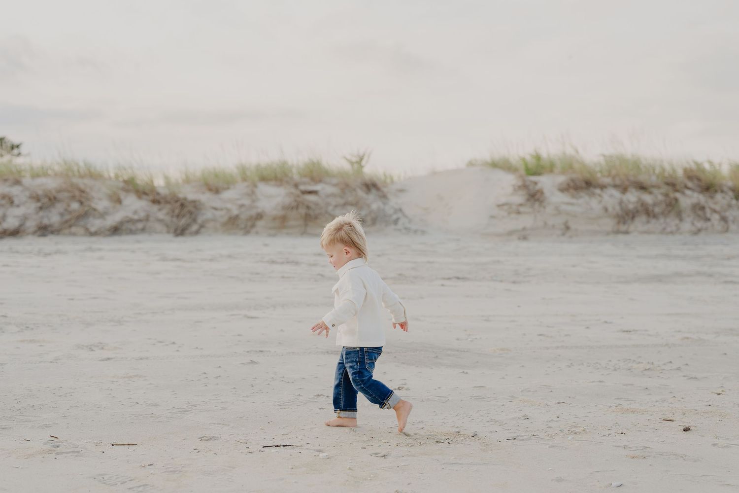 A young toddler with blonde hair runs barefoot on a sandy beach wearing jeans and a white top.