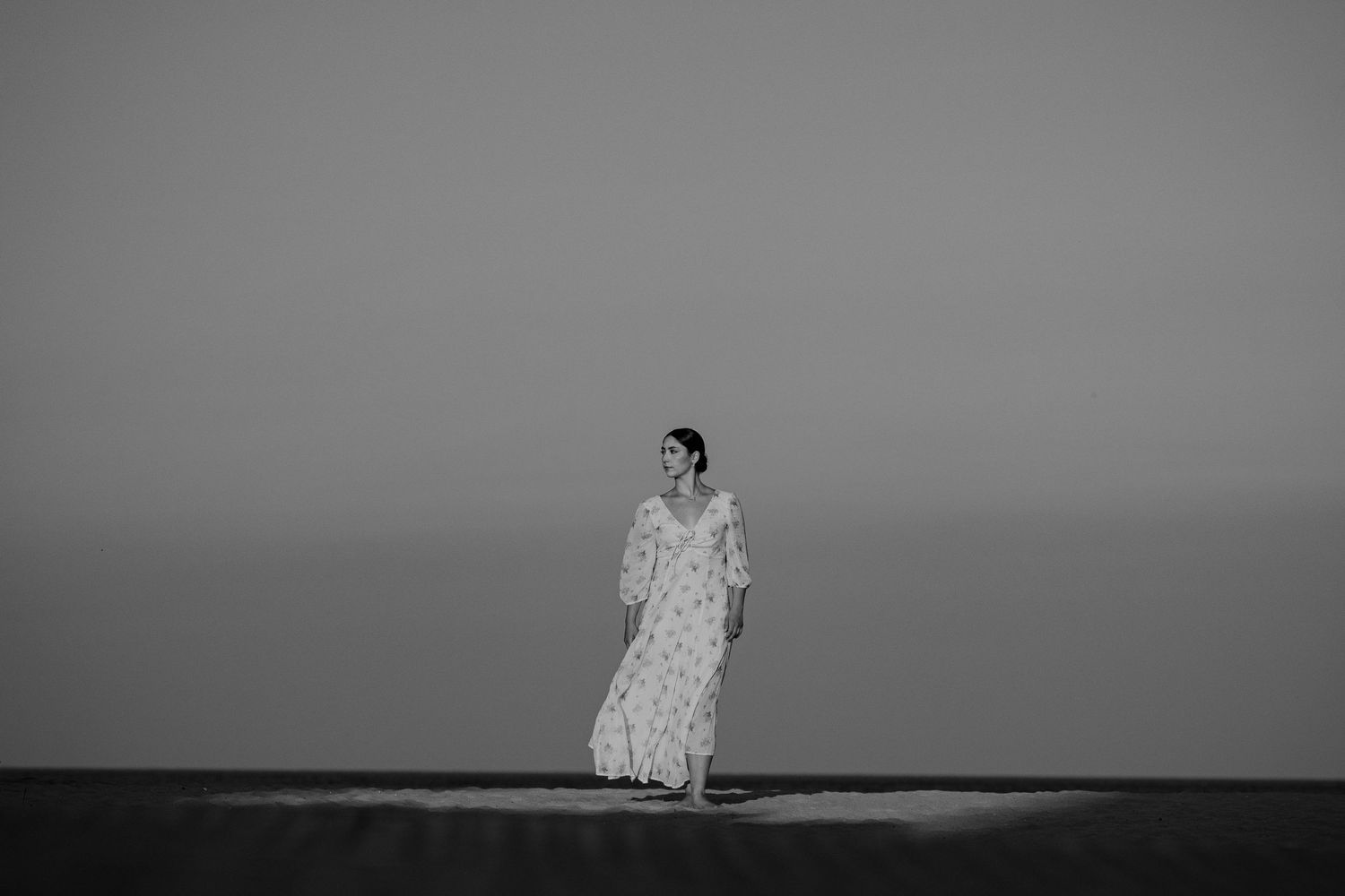 Black and white photo of a woman in a floral maxi dress standing alone on a beach at dusk.