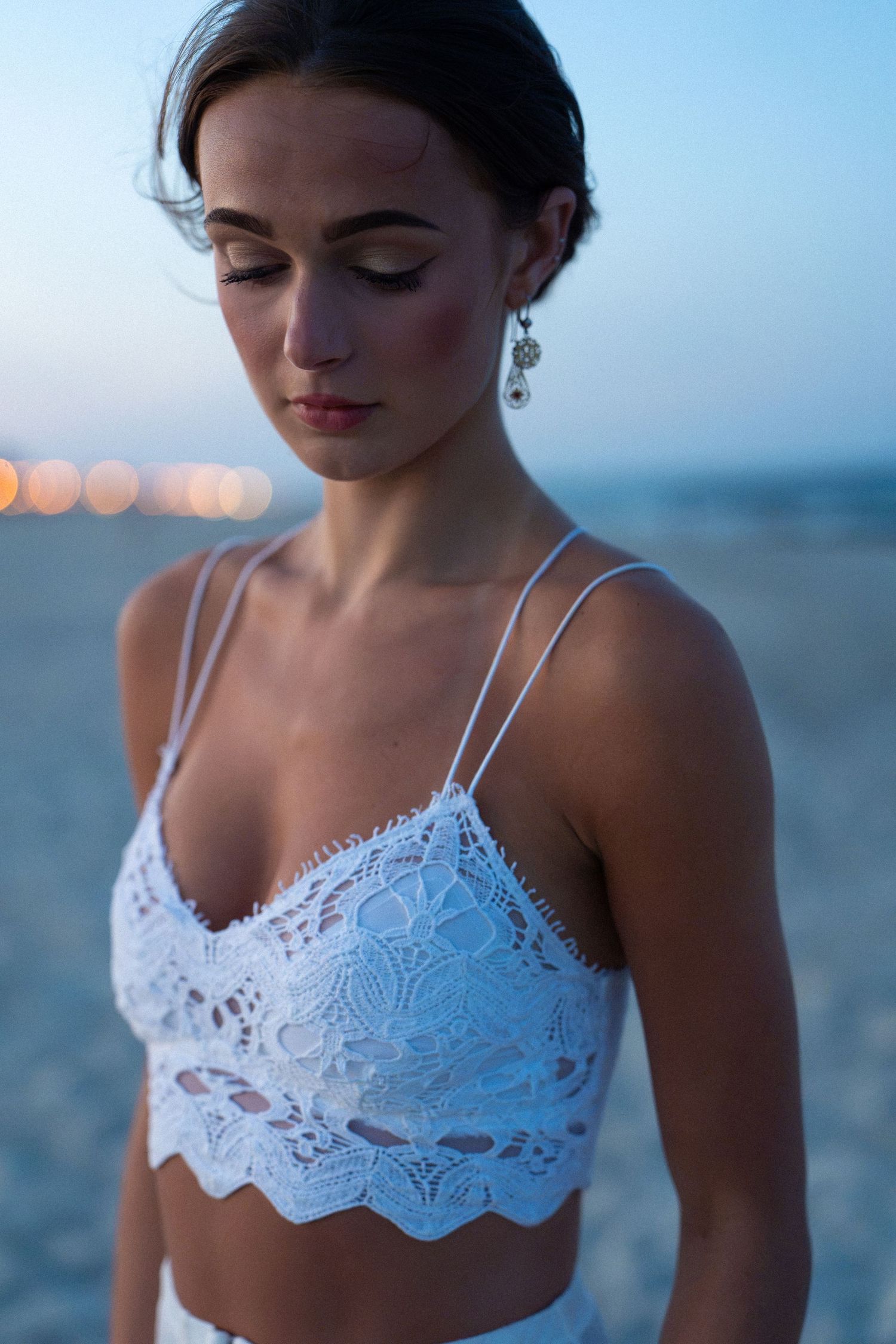 Woman in white lace bralette with dangling earrings poses on beach at twilight.
