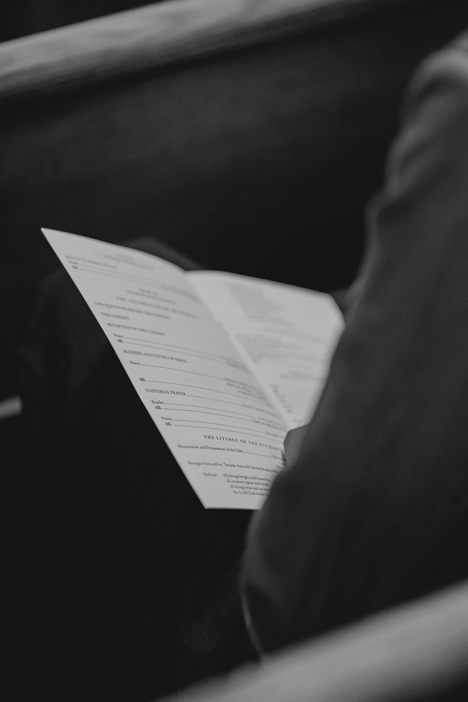 Black and white photo of a person reading an open book or program while seated in a dark wooden pew.