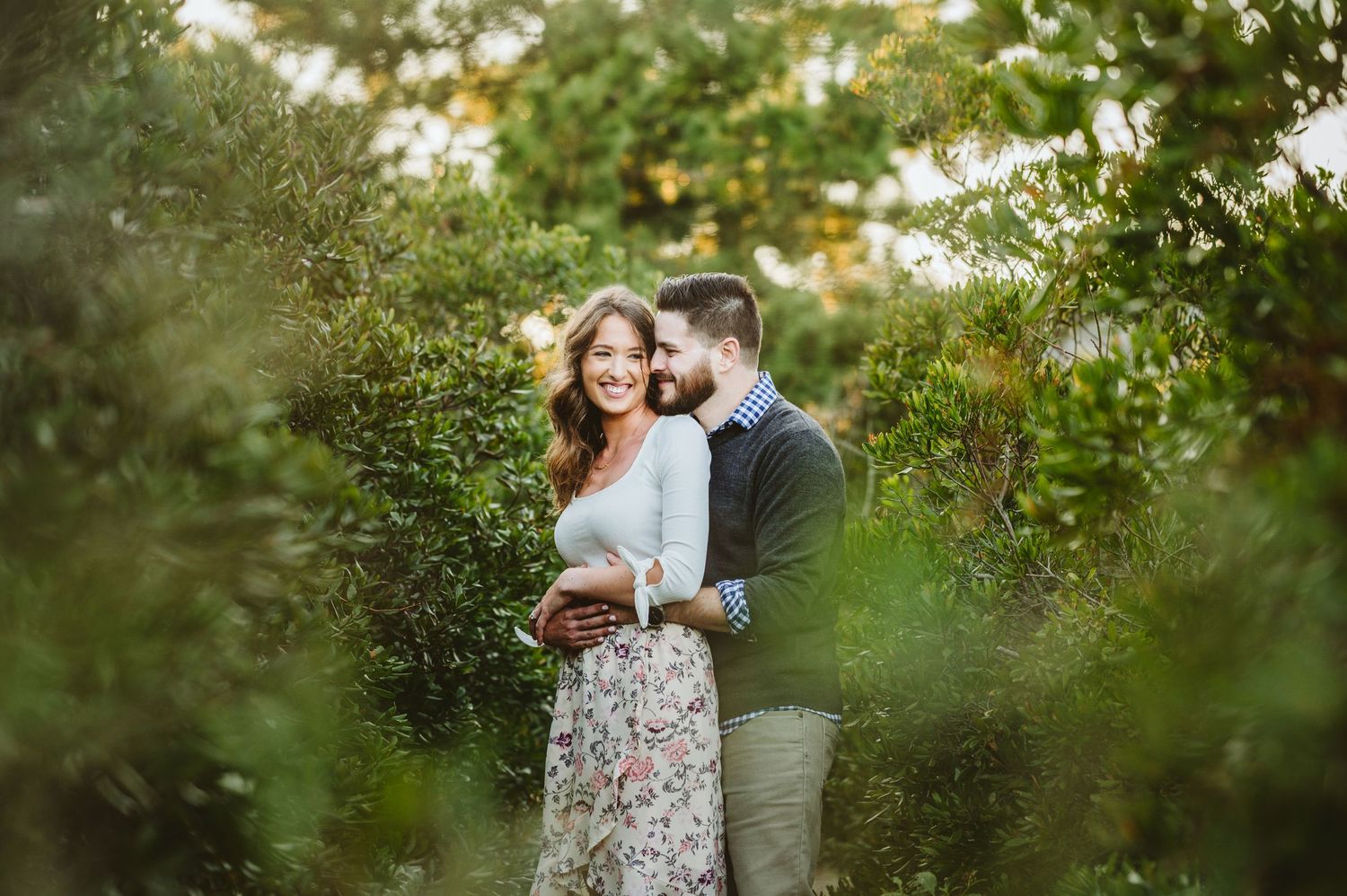 Romantic engagement photos of couple in casual attire among lush greenery.