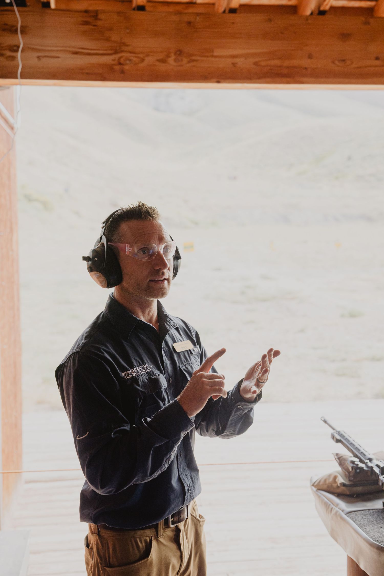 Person in black jacket gestures while wearing protective headphones in a workshop setting.