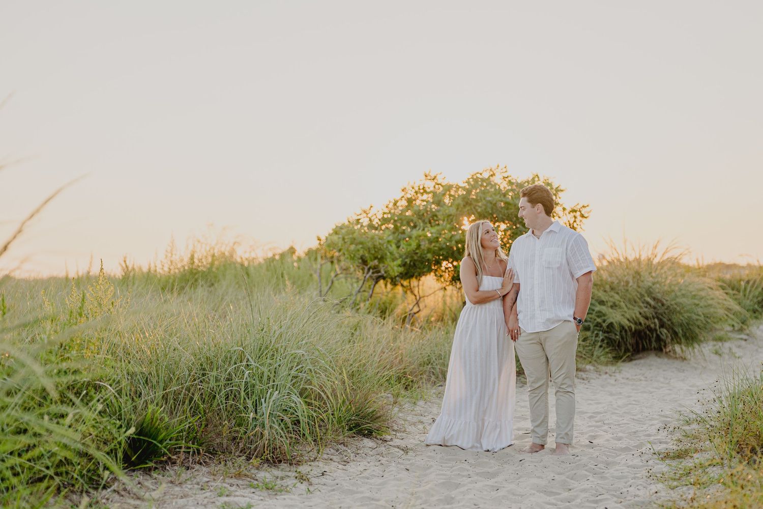 Couple holding hands on sandy beach path surrounded by tall grass at golden hour sunset.