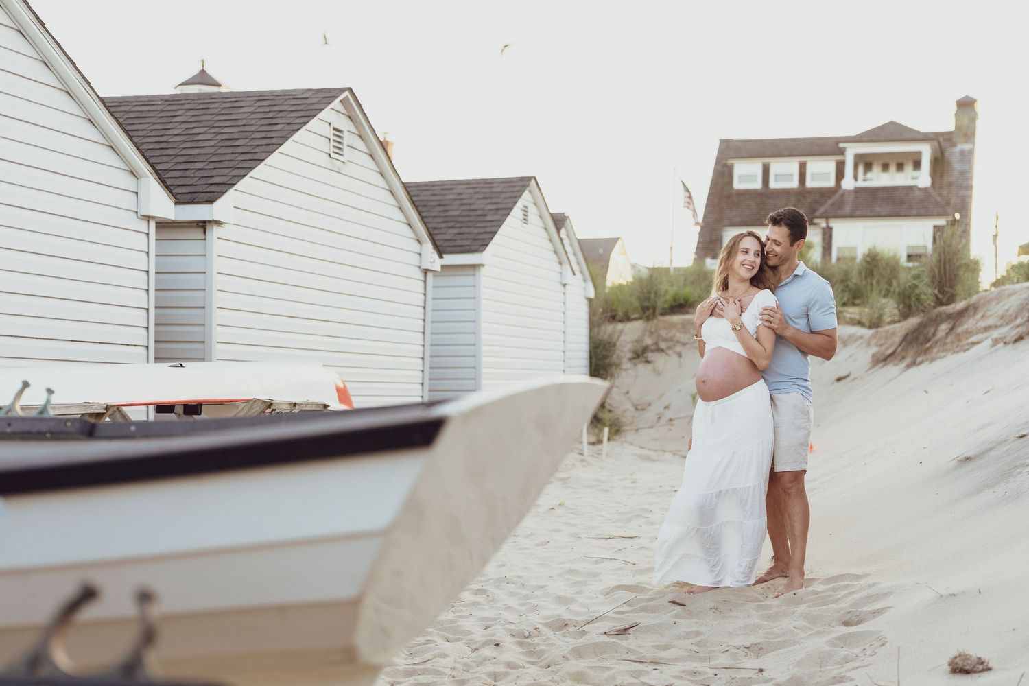 Beach houses and boats line the shore as a couple walks along the sand.