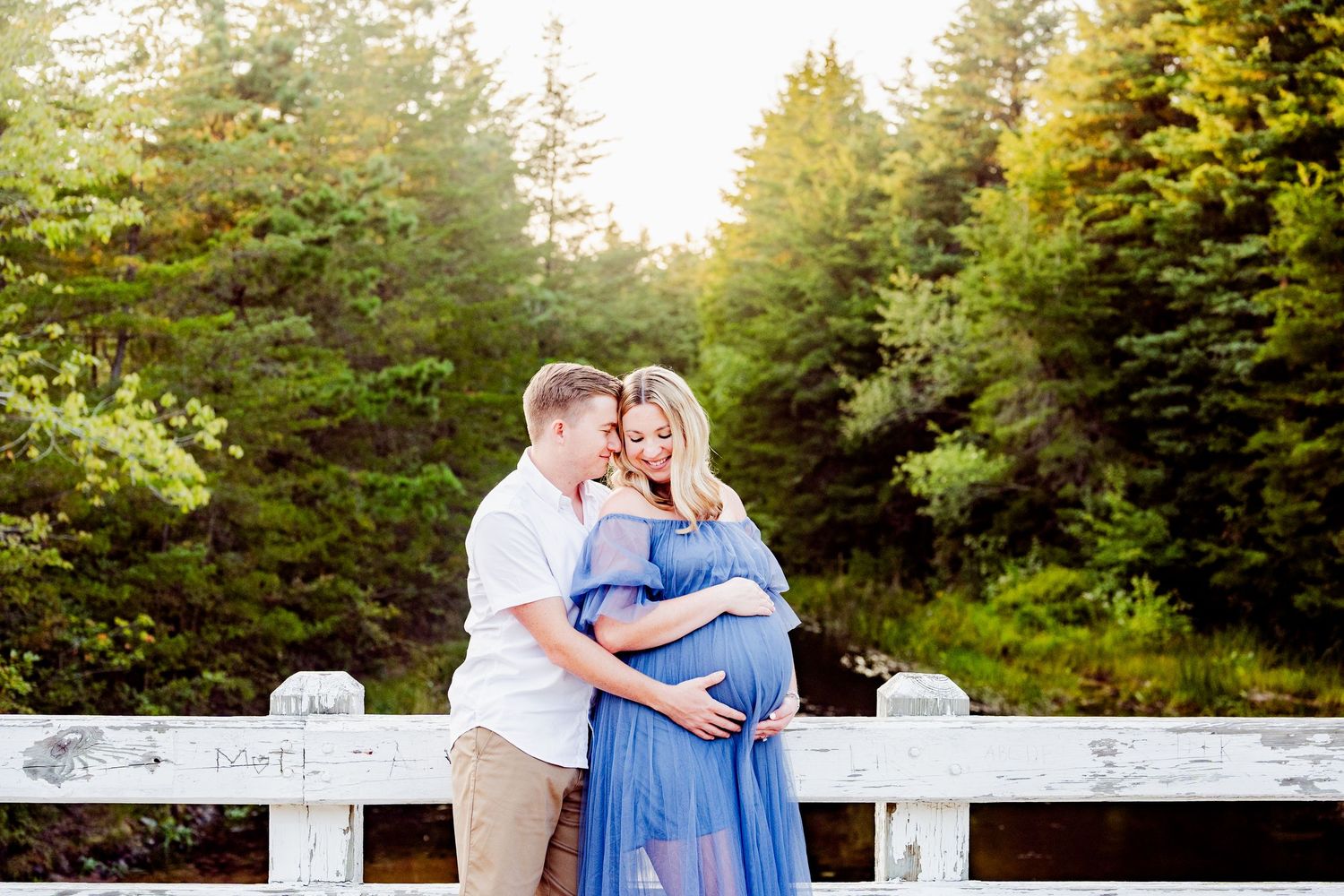 Maternity photo of a couple sharing an intimate moment on a white bridge in a forest setting.