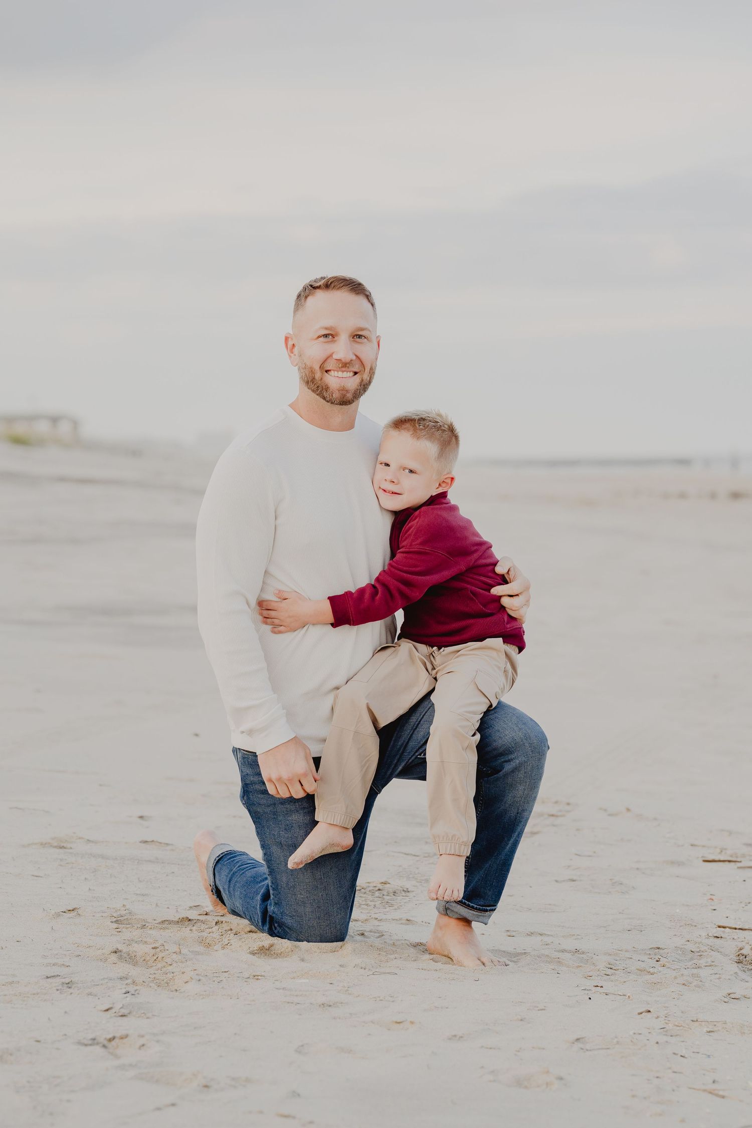 A smiling father kneels on a sandy beach hugging his young son in a red sweater on an overcast day.