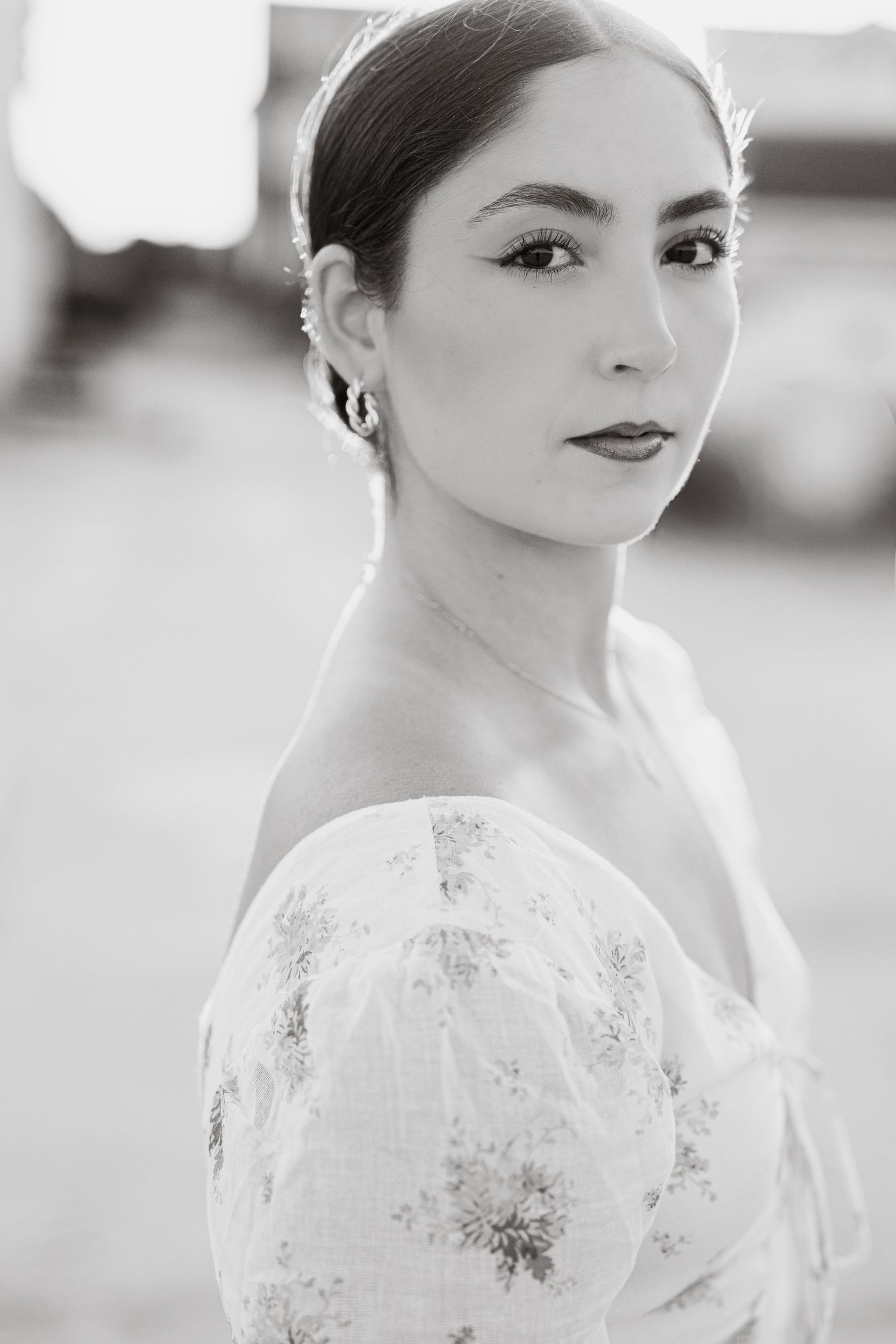 Black and white portrait of a young woman in an off-shoulder floral dress with hoop earrings, gazing over her shoulder.