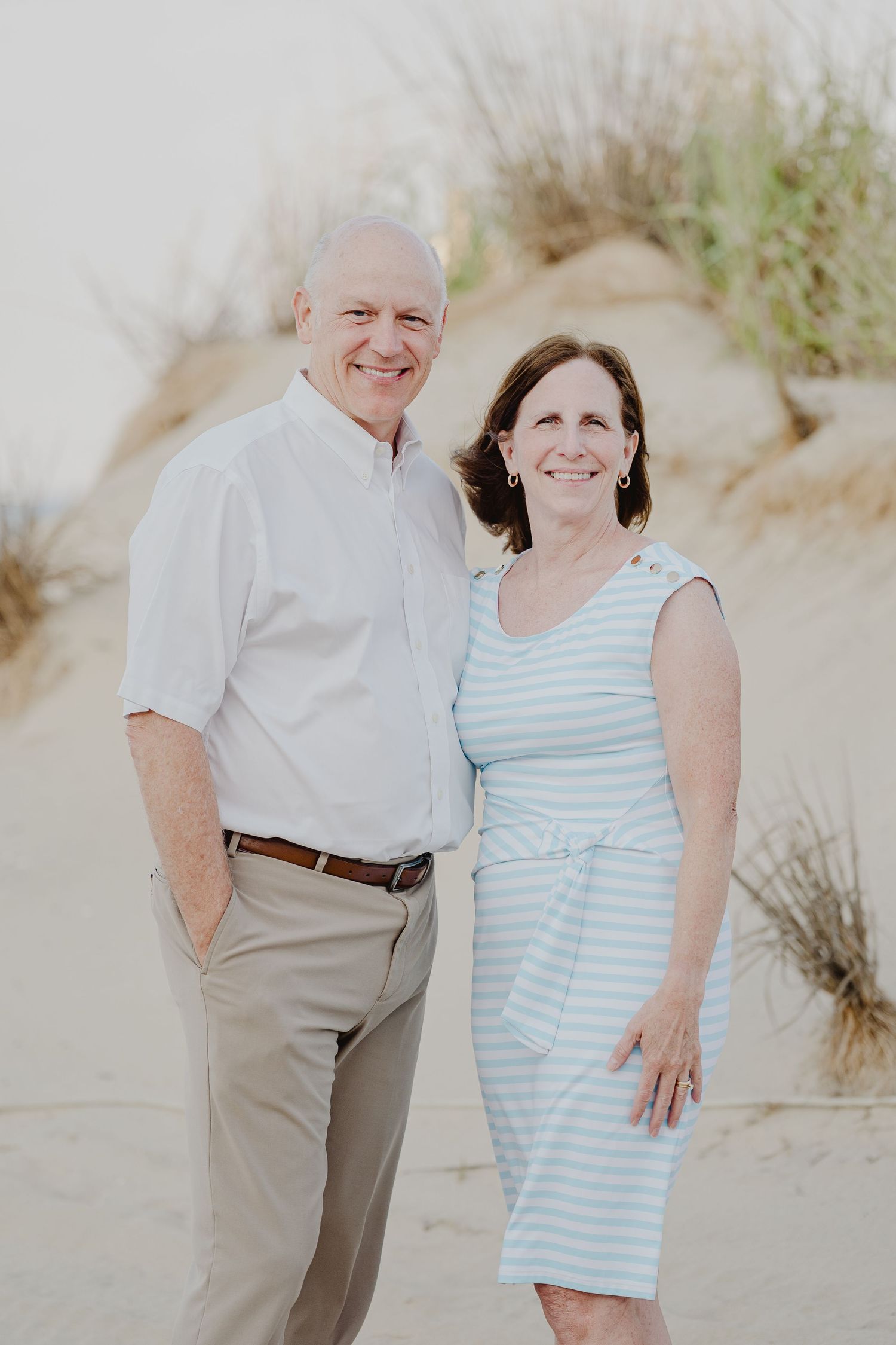 A smiling couple poses together on a sandy beach, man in white shirt and woman in light blue striped dress.