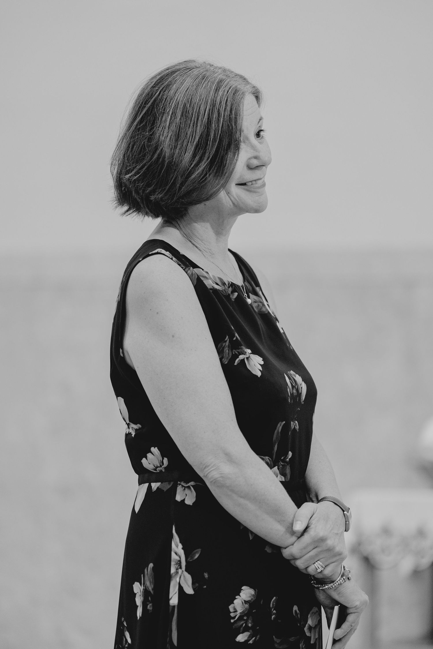 Black and white portrait of a smiling middle-aged woman in a floral dress standing outdoors.
