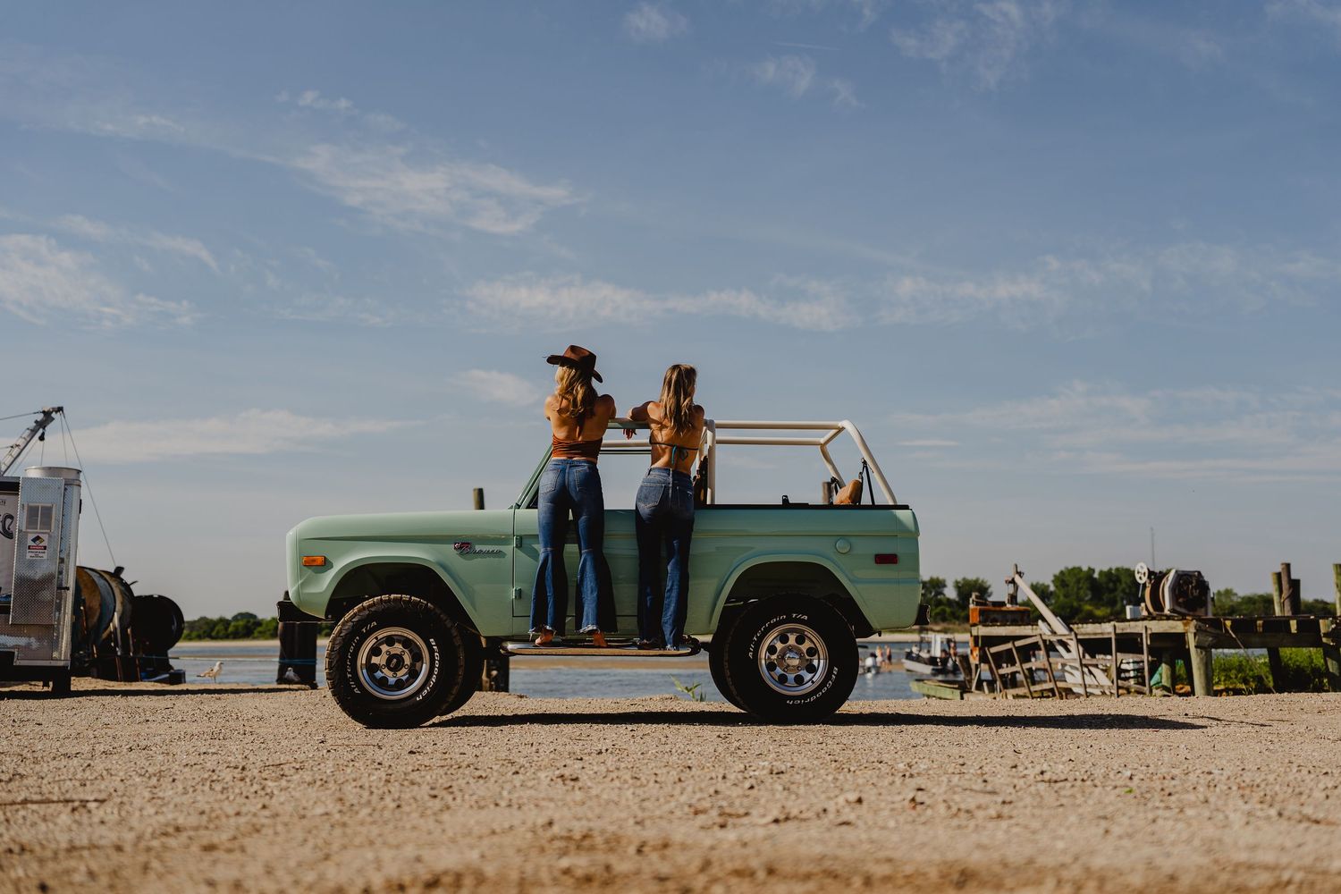 Two women in cowboy hats posing atop a classic mint green Ford Bronco on a gravel lot.