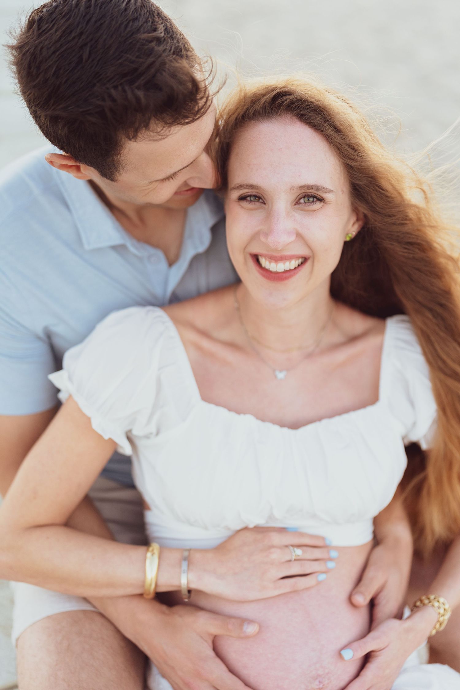 A couple in white and blue clothing embraces during a beachside maternity session.