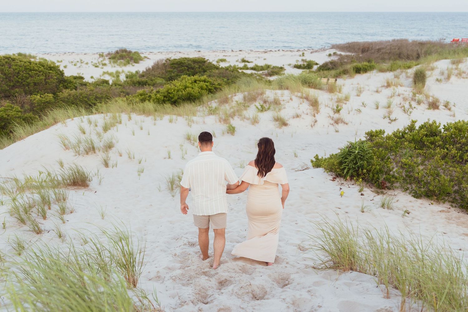 A couple walks hand in hand down sandy dunes with ocean views.