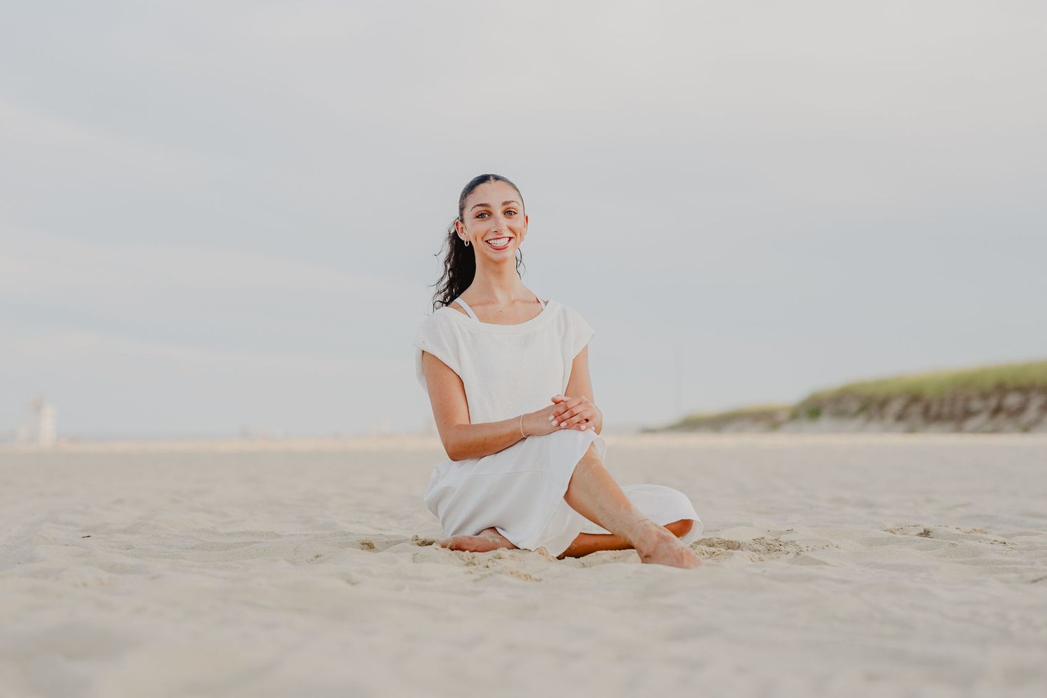 Smiling woman in white dress sitting cross-legged on sandy beach with dunes in background.