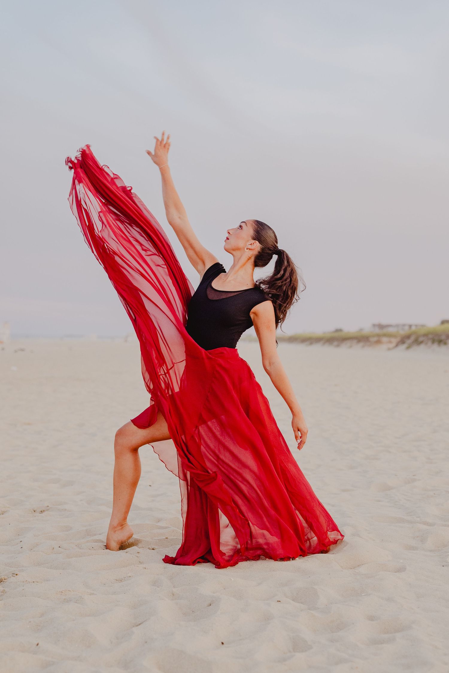 Female dancer in red chiffon skirt and black leotard extends arm upward on sandy beach.