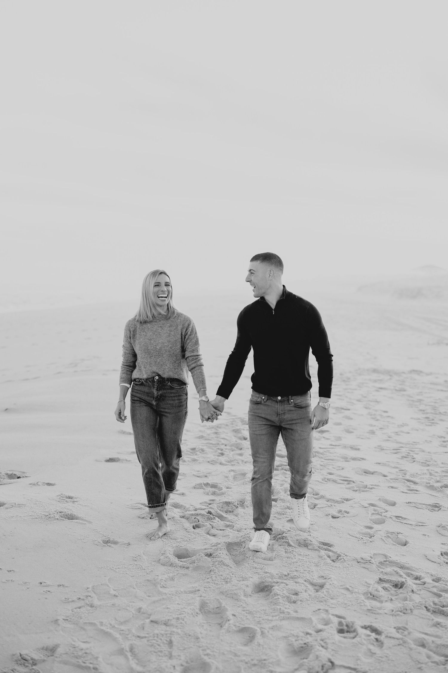 A couple walks hand in hand along a sandy beach in black and white, leaving footprints in their wake.