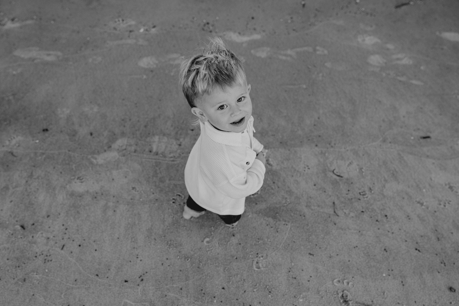 Black and white photo of a happy toddler boy standing on sandy ground, looking up at the camera.