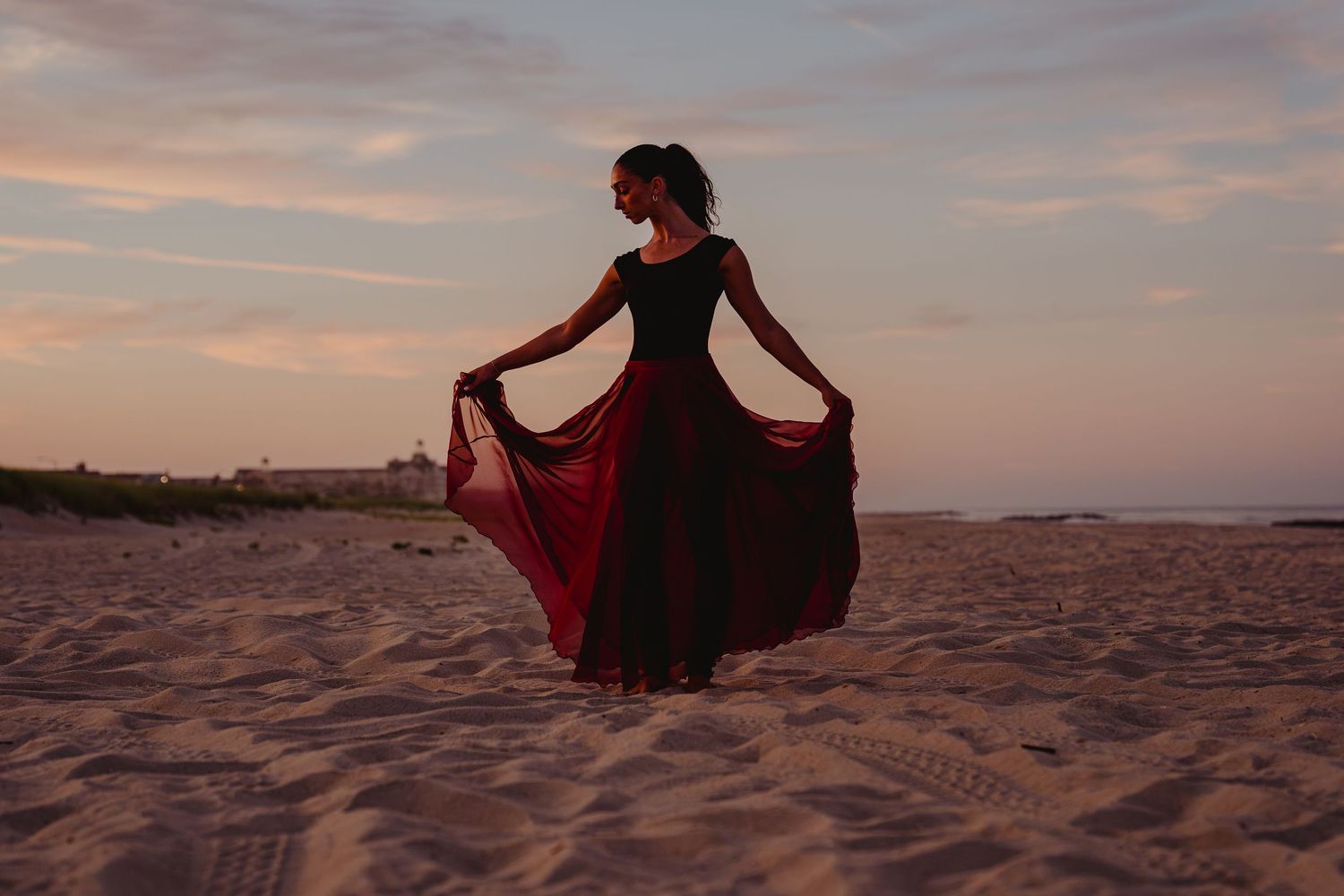 Woman in elegant black and red flowing dress twirling on sandy beach at sunset.