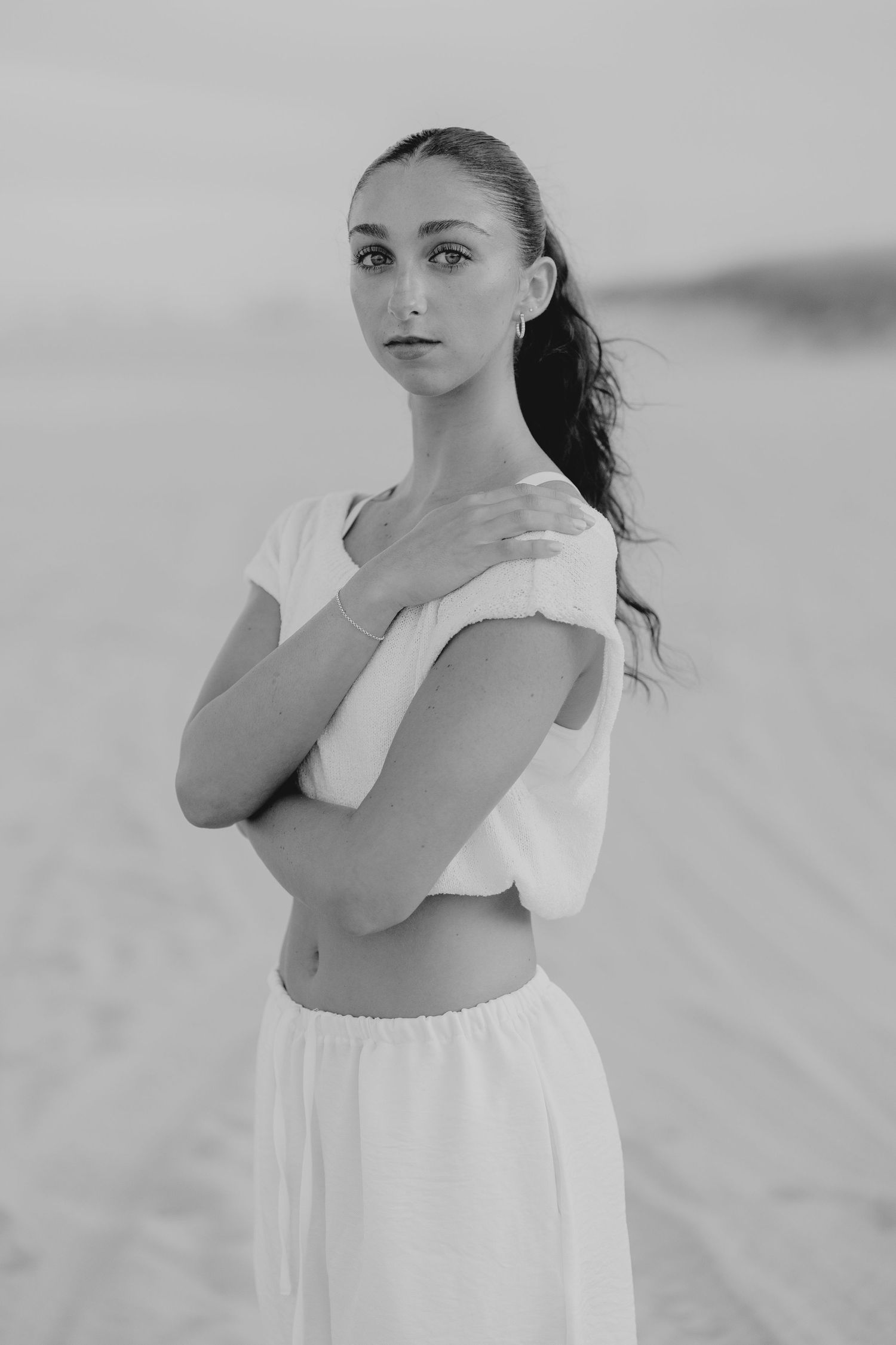 Black and white portrait of a young woman in white crop top and skirt, posing gracefully in a desert landscape.