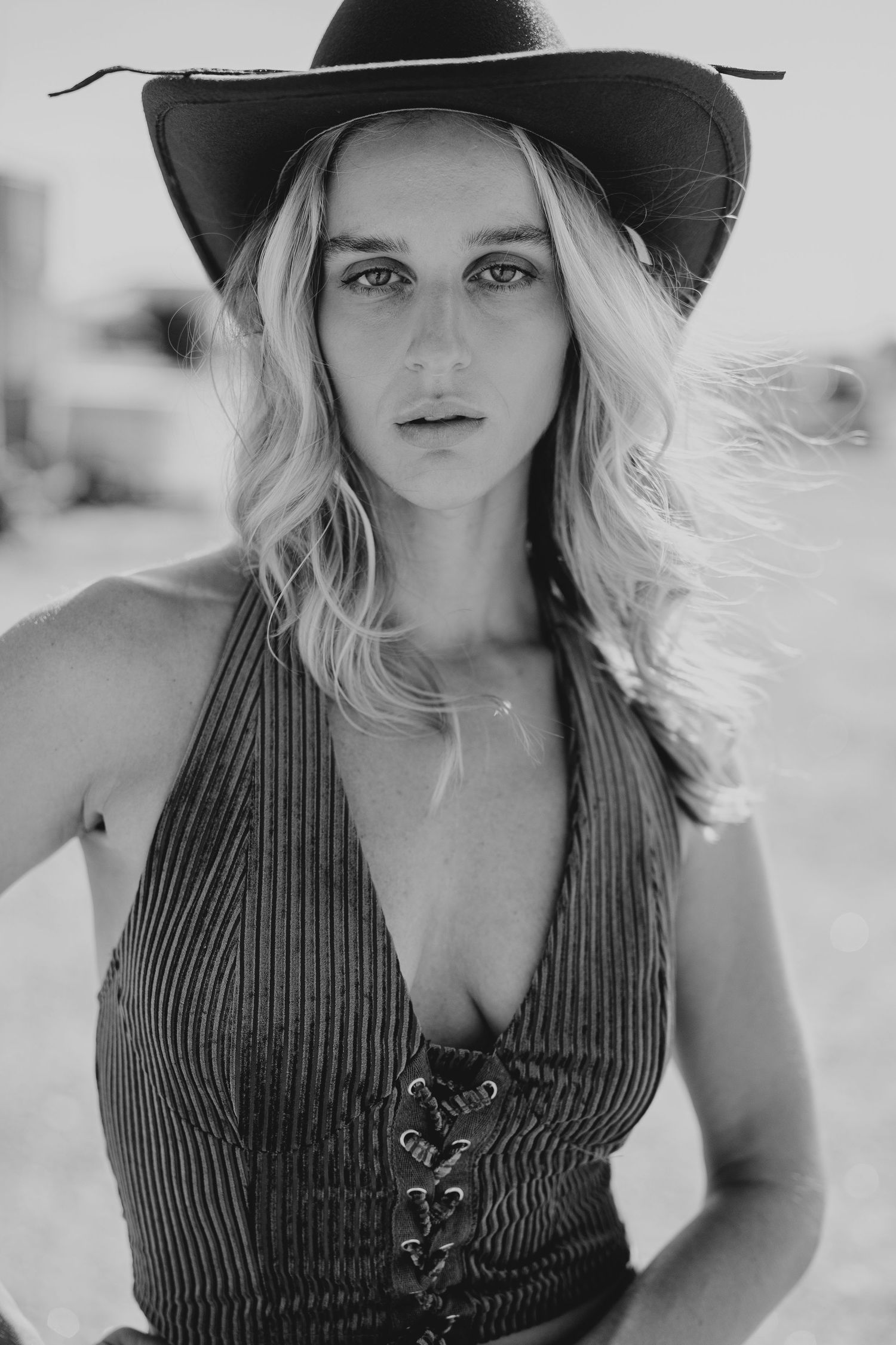 Black and white portrait of a blonde woman wearing a cowboy hat and lace-up top outdoors.