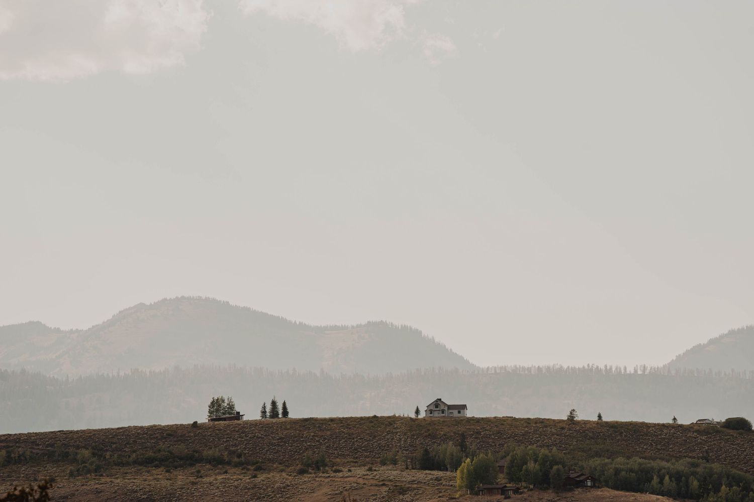 Misty mountain landscape with silhouetted trees and buildings on a hilltop against a moody gray sky.
