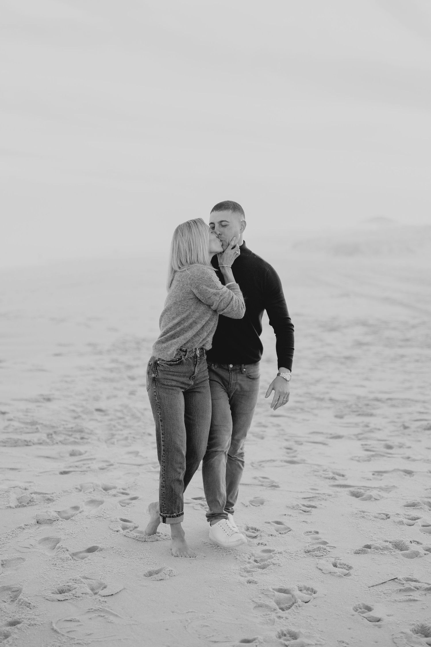 A couple shares an intimate moment while walking together on a sandy beach in black and white photography.