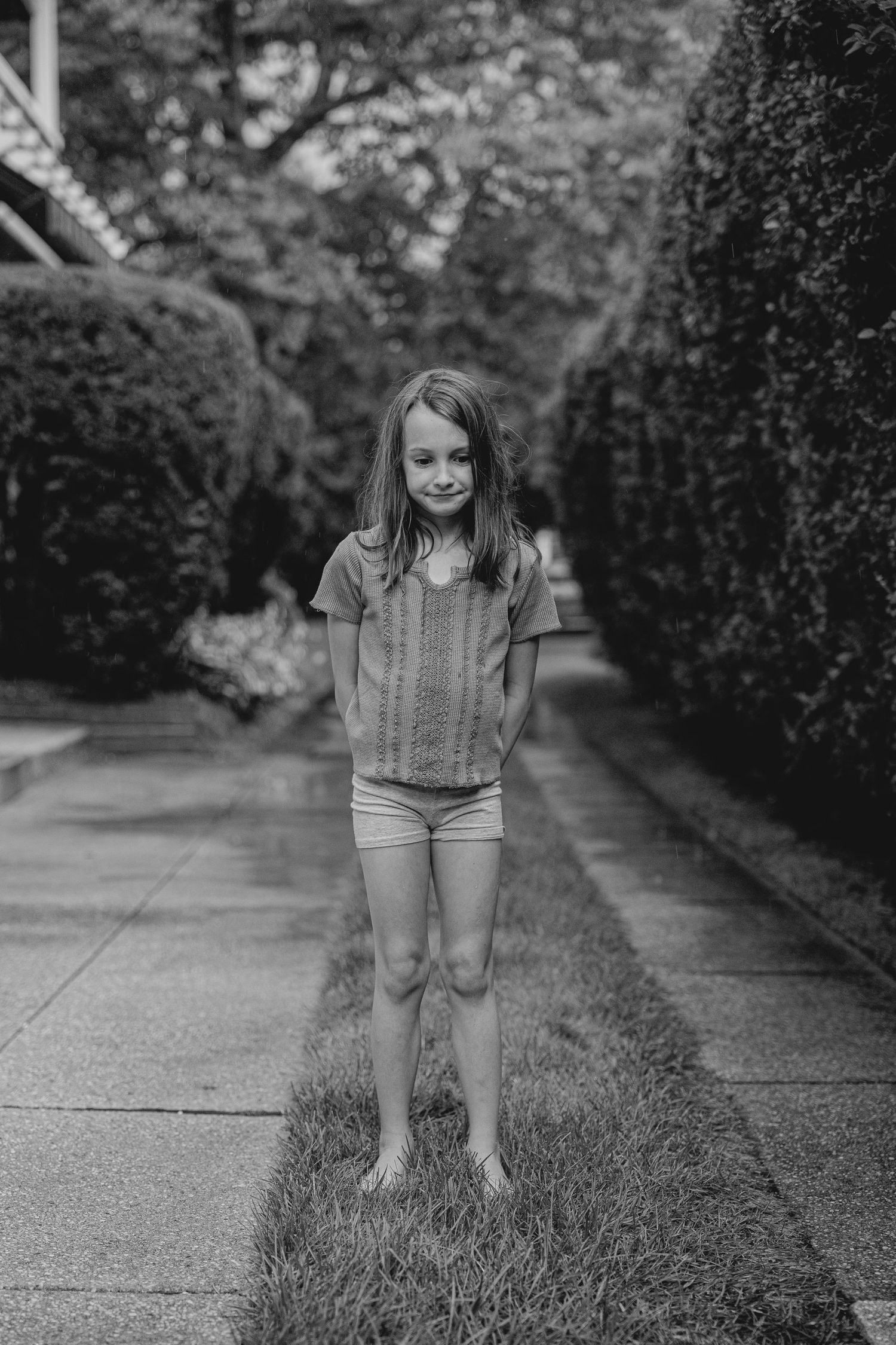 A young barefoot girl in a knit top and shorts stands smiling on a grass path lined with trimmed hedges.