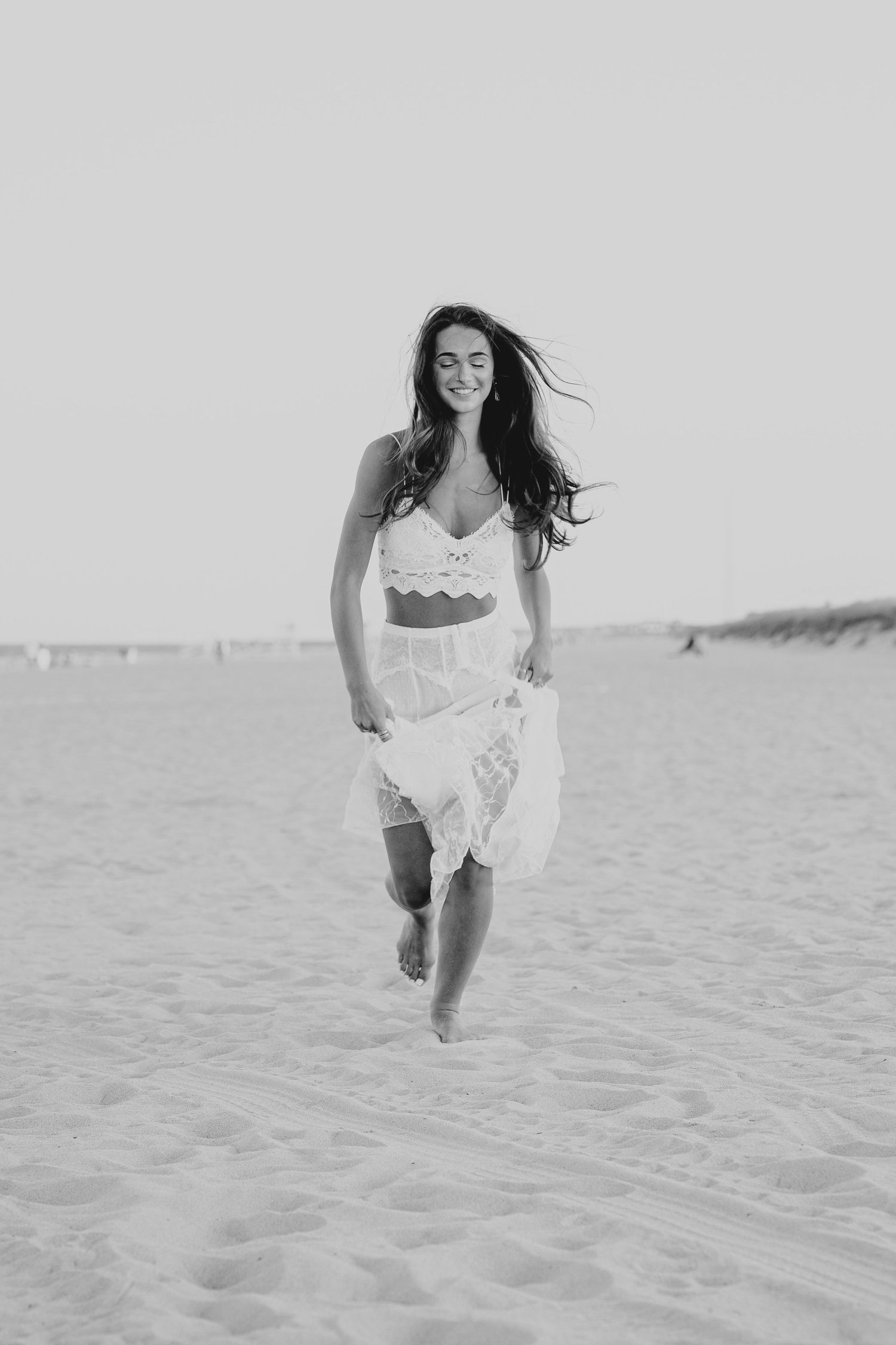Black and white photo of a smiling woman in white crop top and skirt running barefoot on a sandy beach on the Jersey Shore