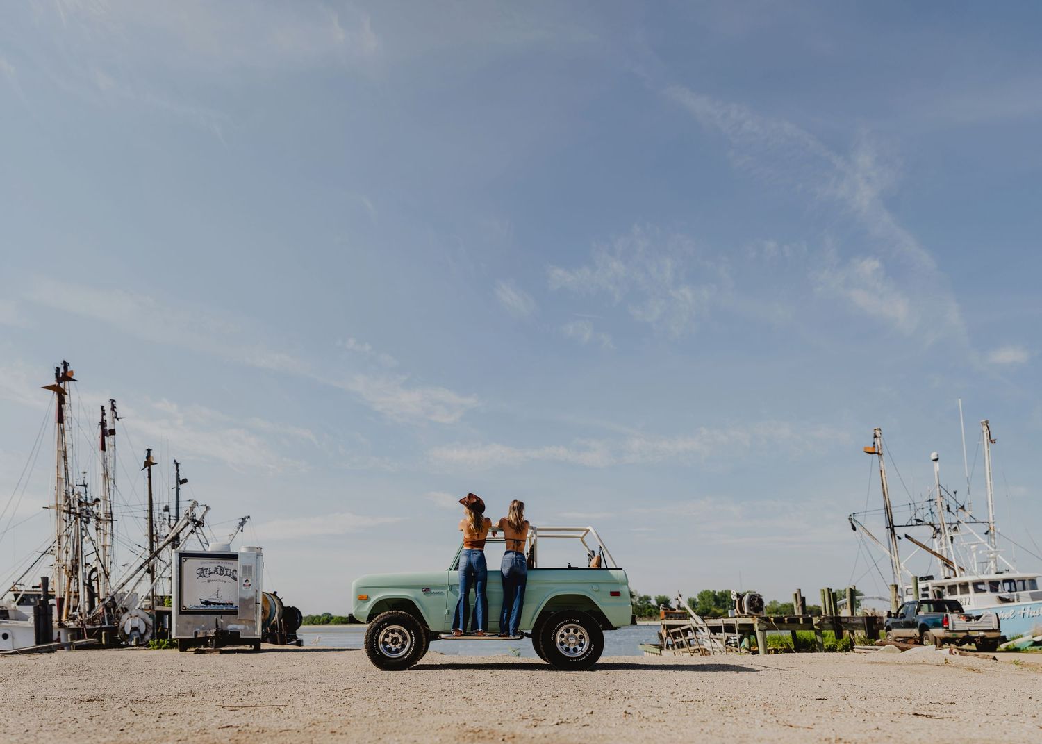 Two women standing on top of a mint green Ford Bronco at a marina with fishing boats in background.