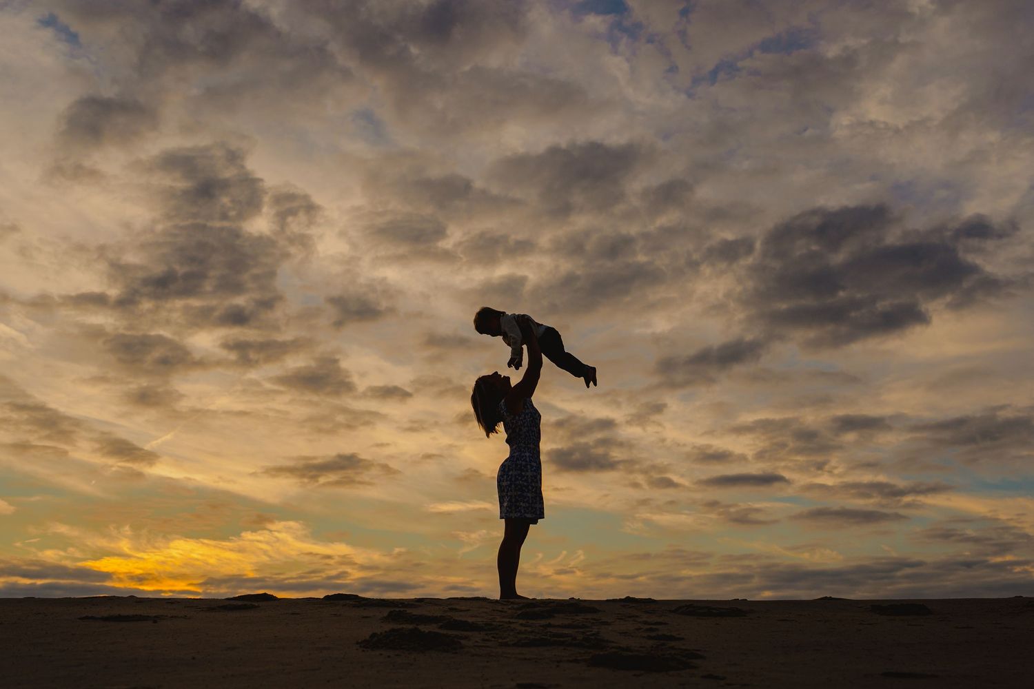 Silhouette of parent lifting baby against dramatic sunset sky with golden clouds on a sandy beach.