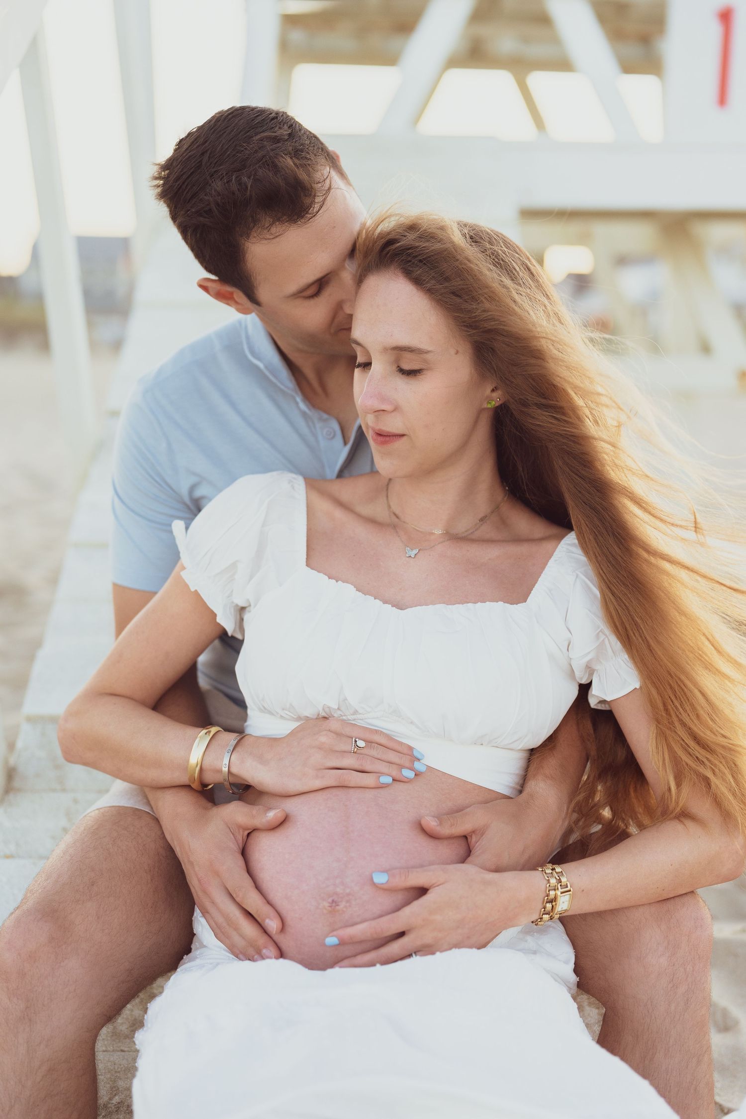 A couple shares tender moments on a beach bench during a maternity photoshoot.