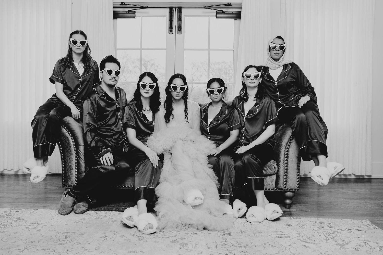 Wedding party in black attire sitting against white wall for stylish group photo.