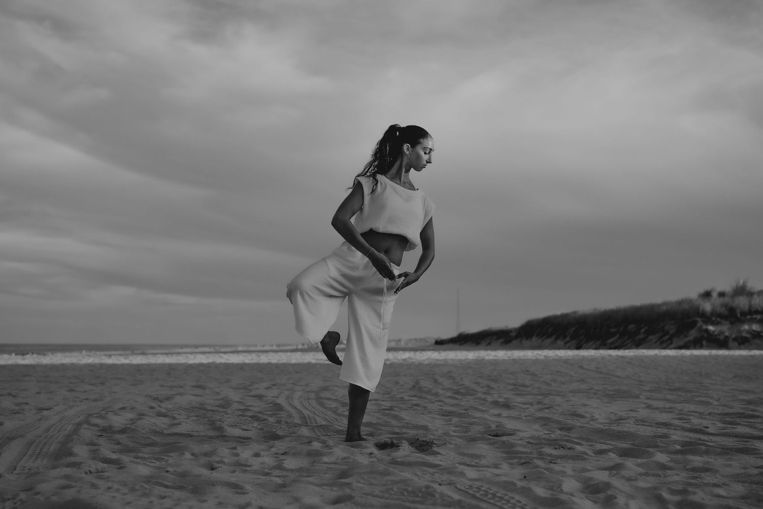 Woman in white outfit dancing on a sandy beach under a cloudy sky in black and white photography.