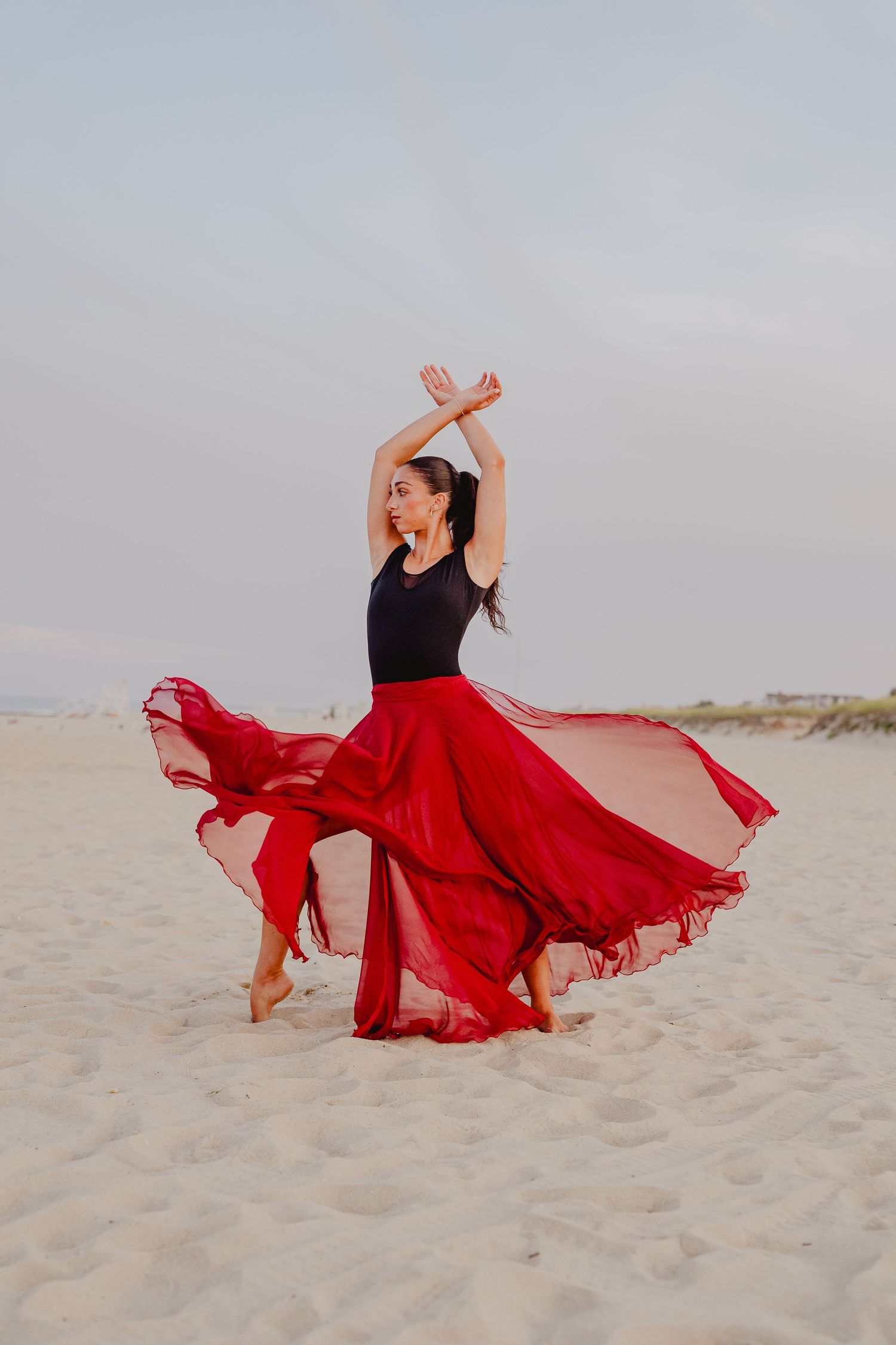 Dancer in black leotard and flowing red skirt performs graceful ballet pose on sandy beach.