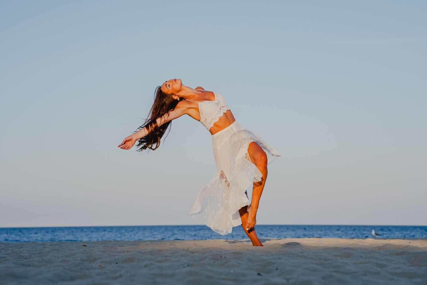Woman in white dress dancing gracefully on a sandy beach at sunset, hair flowing in the breeze.