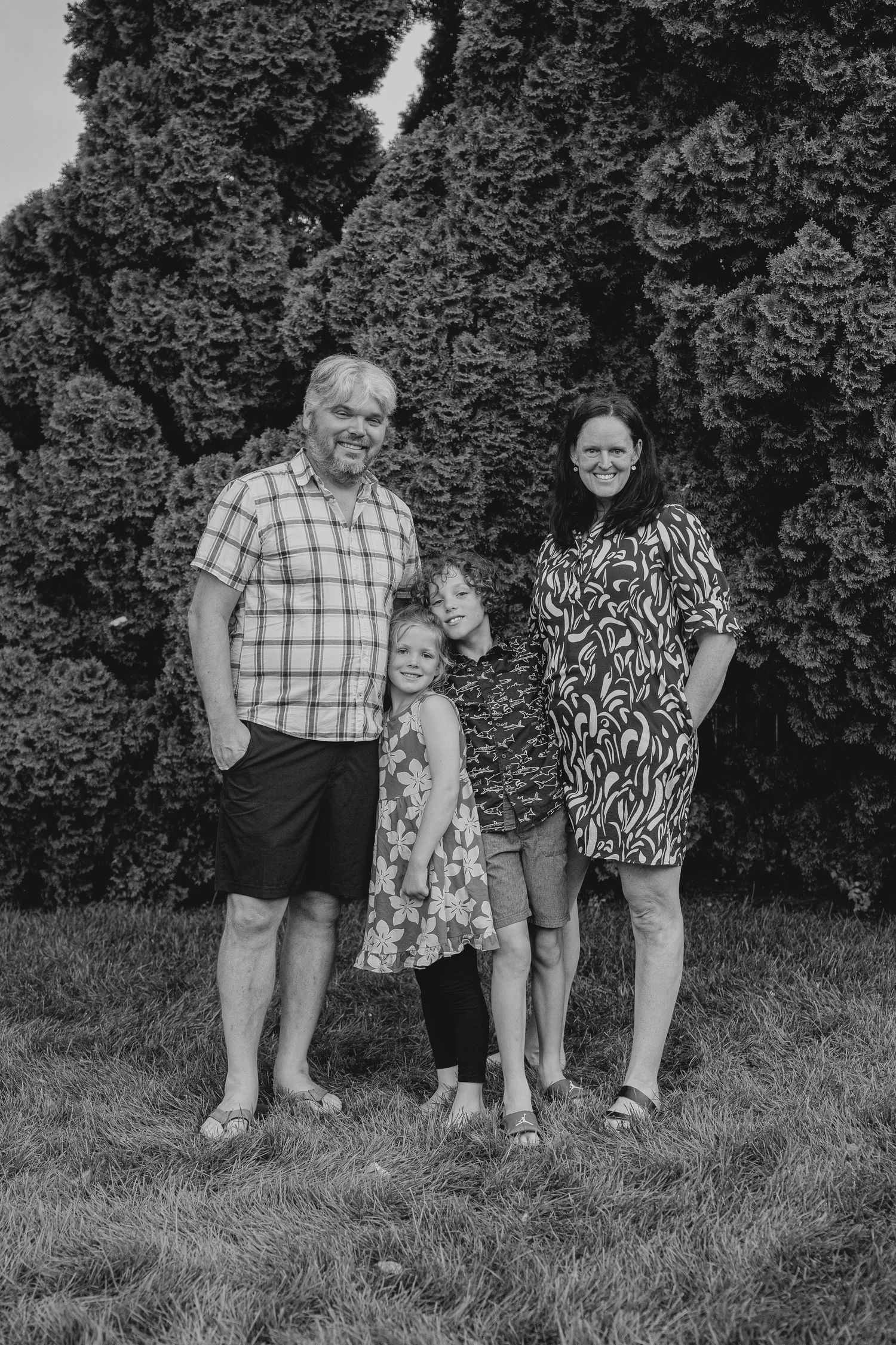 Black and white family portrait of two adults and two children smiling outdoors on grass with large evergreen trees behind them.