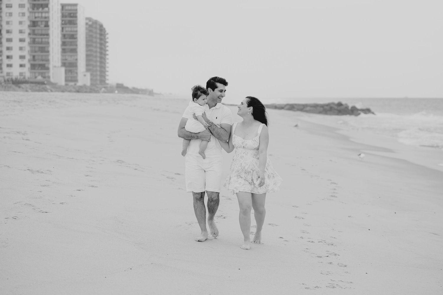 Black and white photo of a young family walking barefoot on a beach, man holding baby while woman walks beside them.