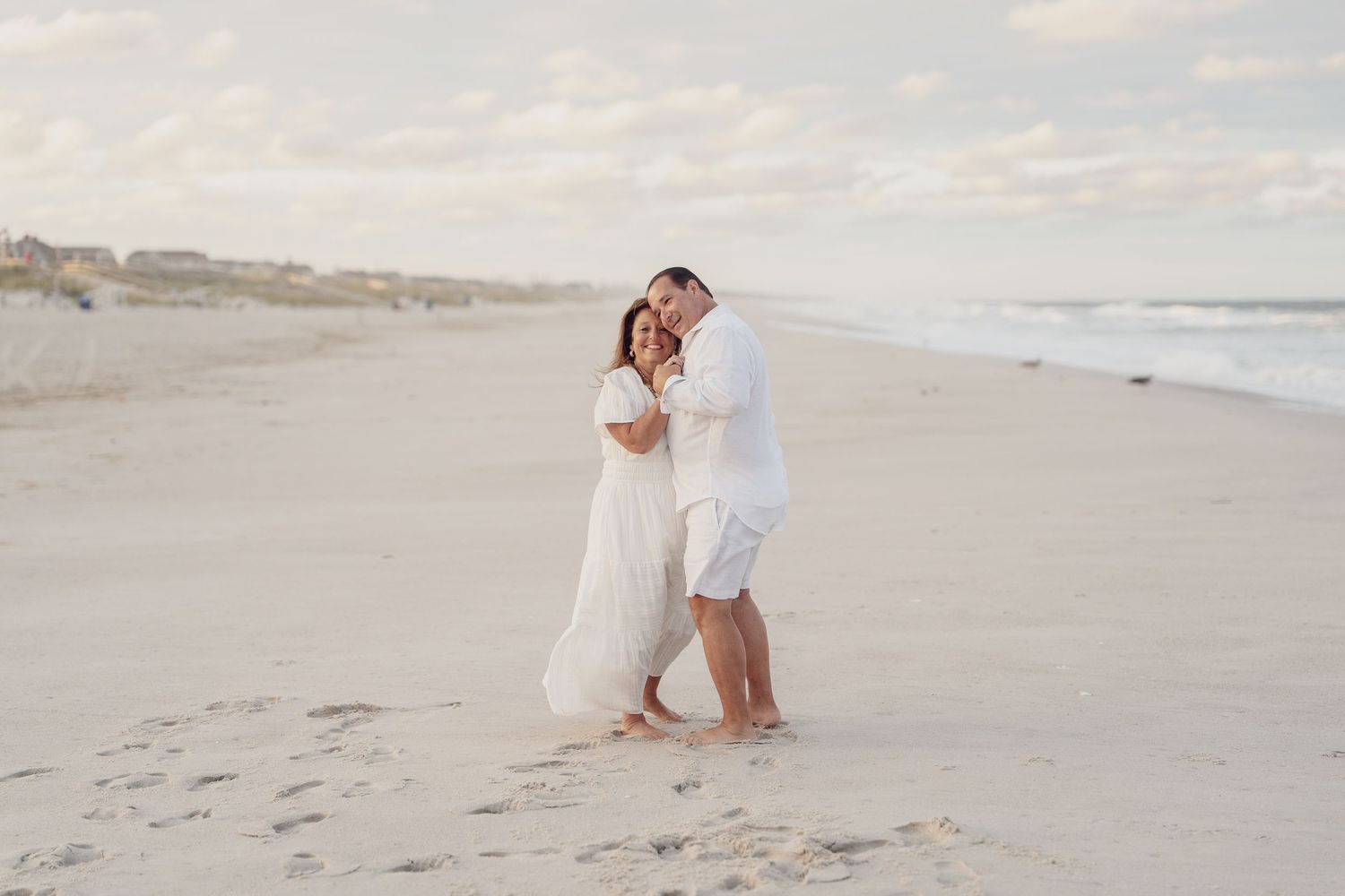 Couple in white outfits walking together along a serene beach shoreline at sunset.