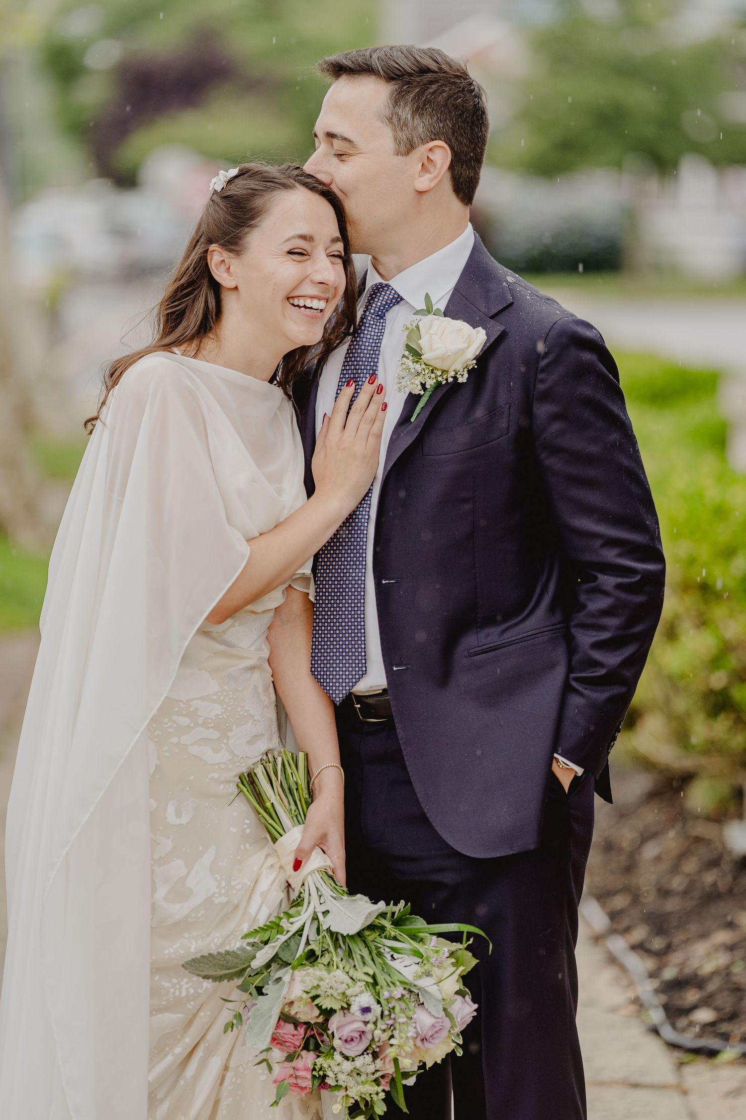 Joyful bride laughing as groom whispers in her ear outdoors, bride holding a pastel wedding bouquet.