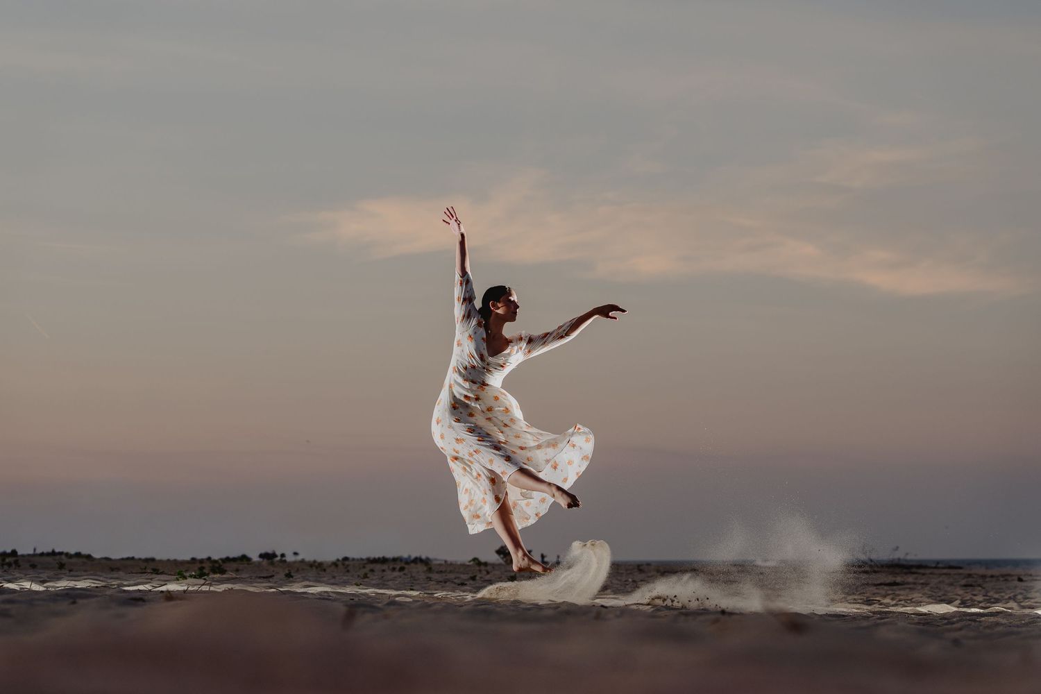 A woman in a flowing white dress leaps gracefully on a beach at sunset, waves crashing behind her.