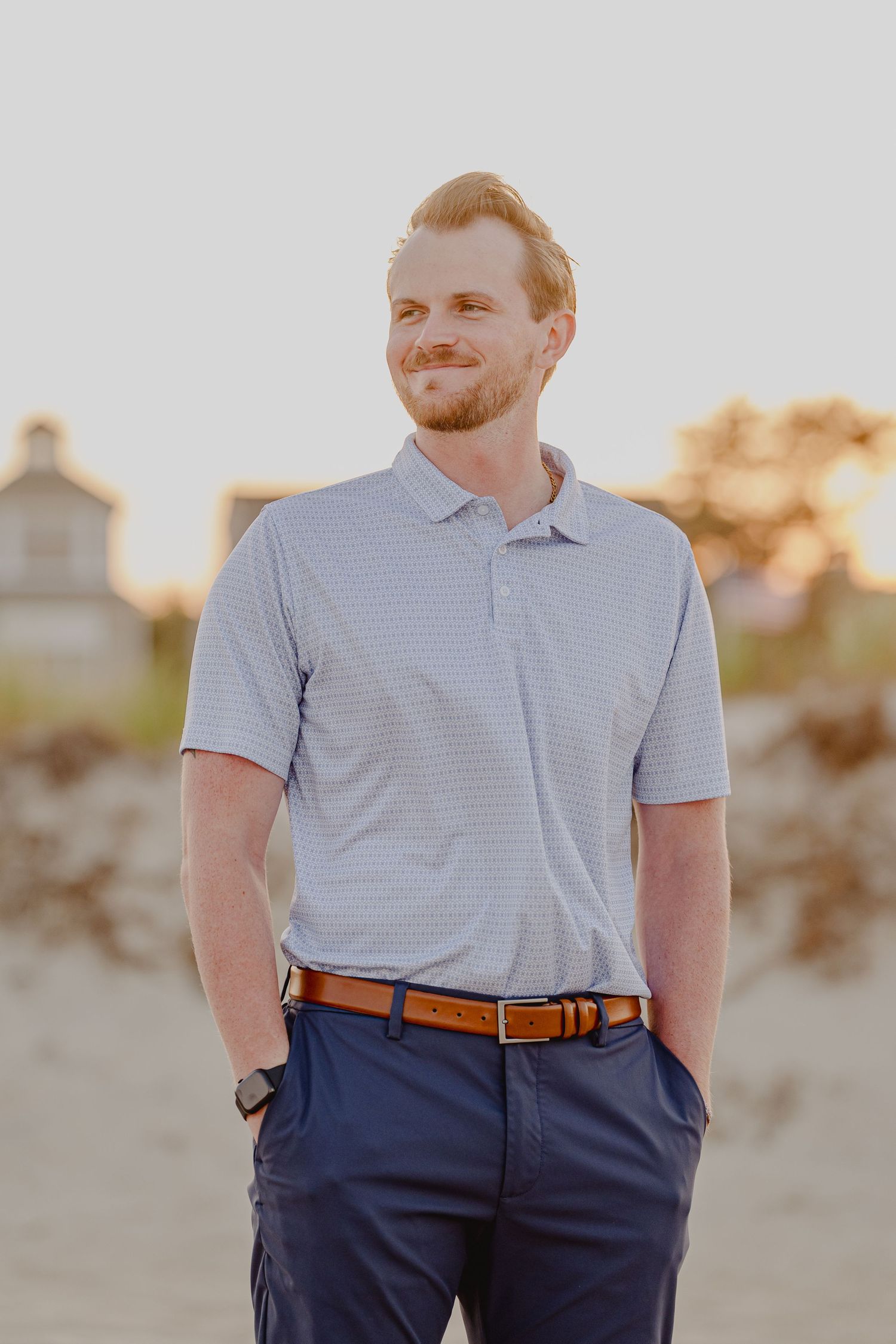 Young man with beard in striped polo shirt and navy pants standing on beach at golden hour sunset.