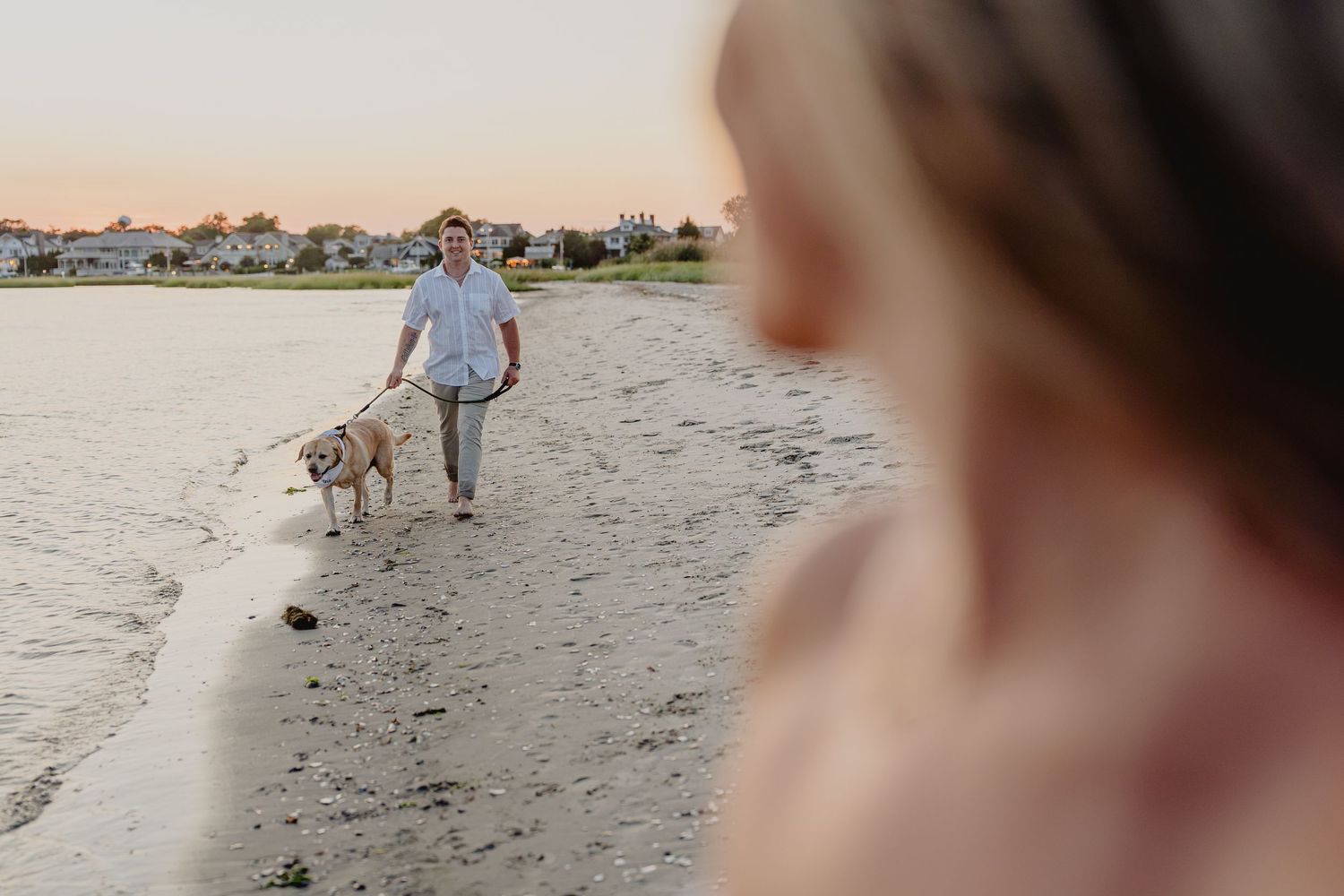 Man walking a yellow Labrador on a sandy beach at sunset with a blurred woman in the foreground.