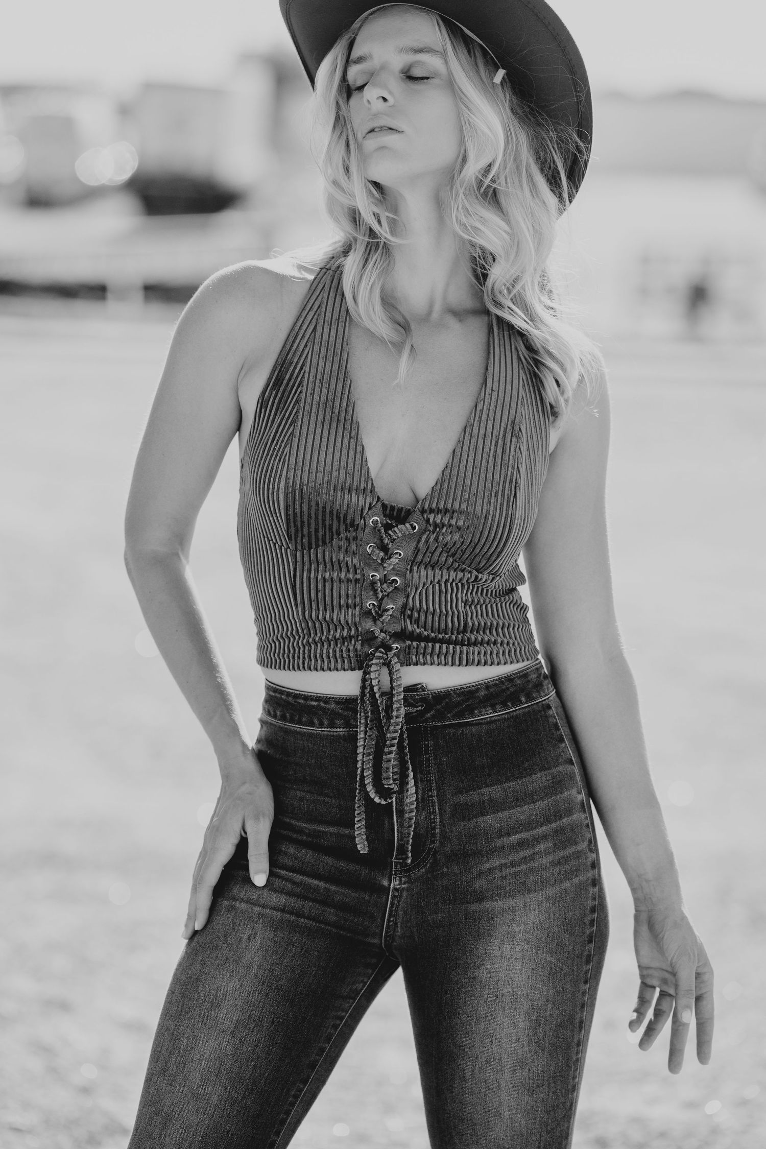 Black and white photo of a blonde woman in a cowboy hat, lace-up crop top, and high-waisted jeans outdoors.