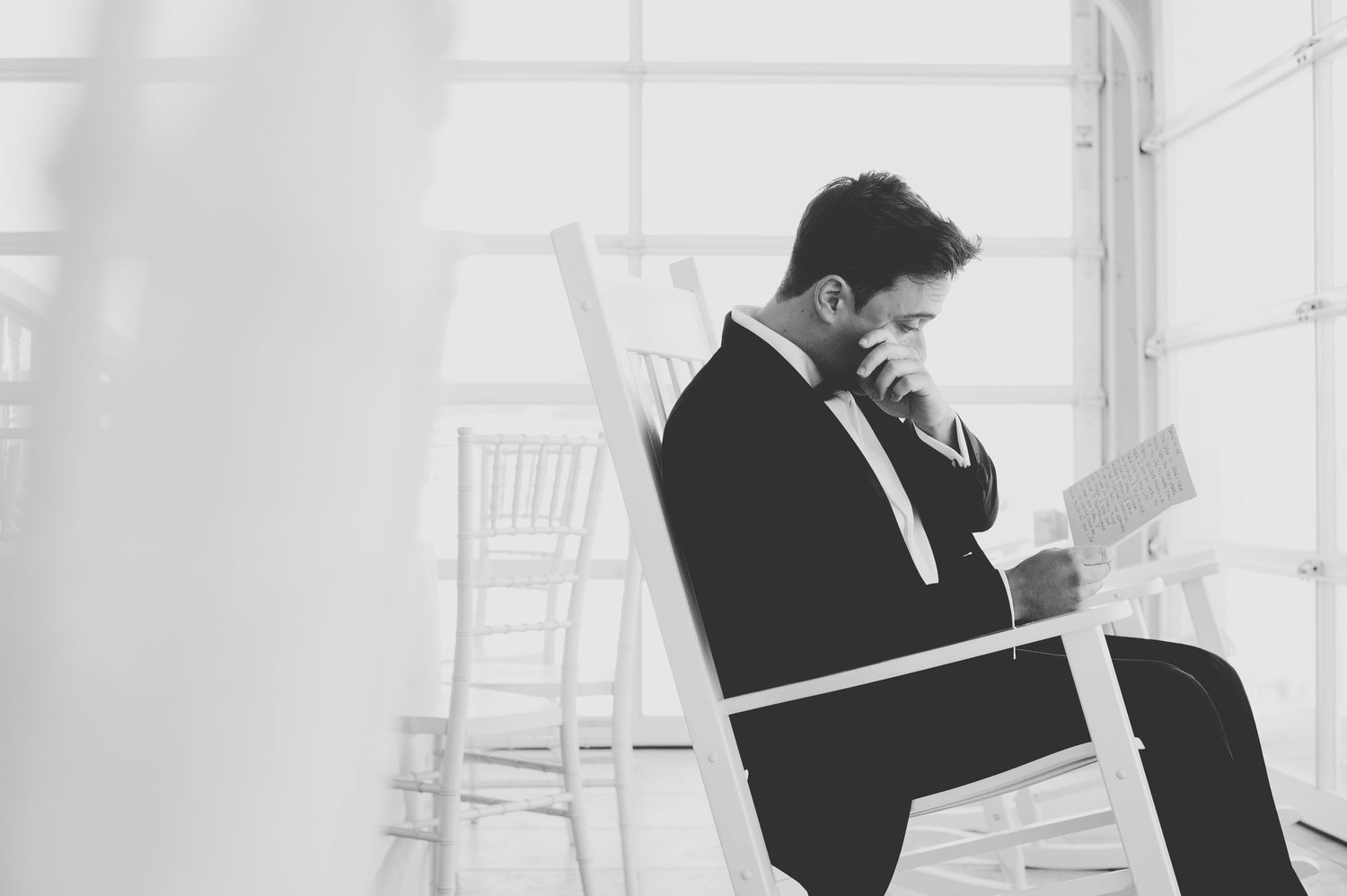 Black and white photo of a groom in a tuxedo sitting on a chair, reading a letter before his wedding ceremony.