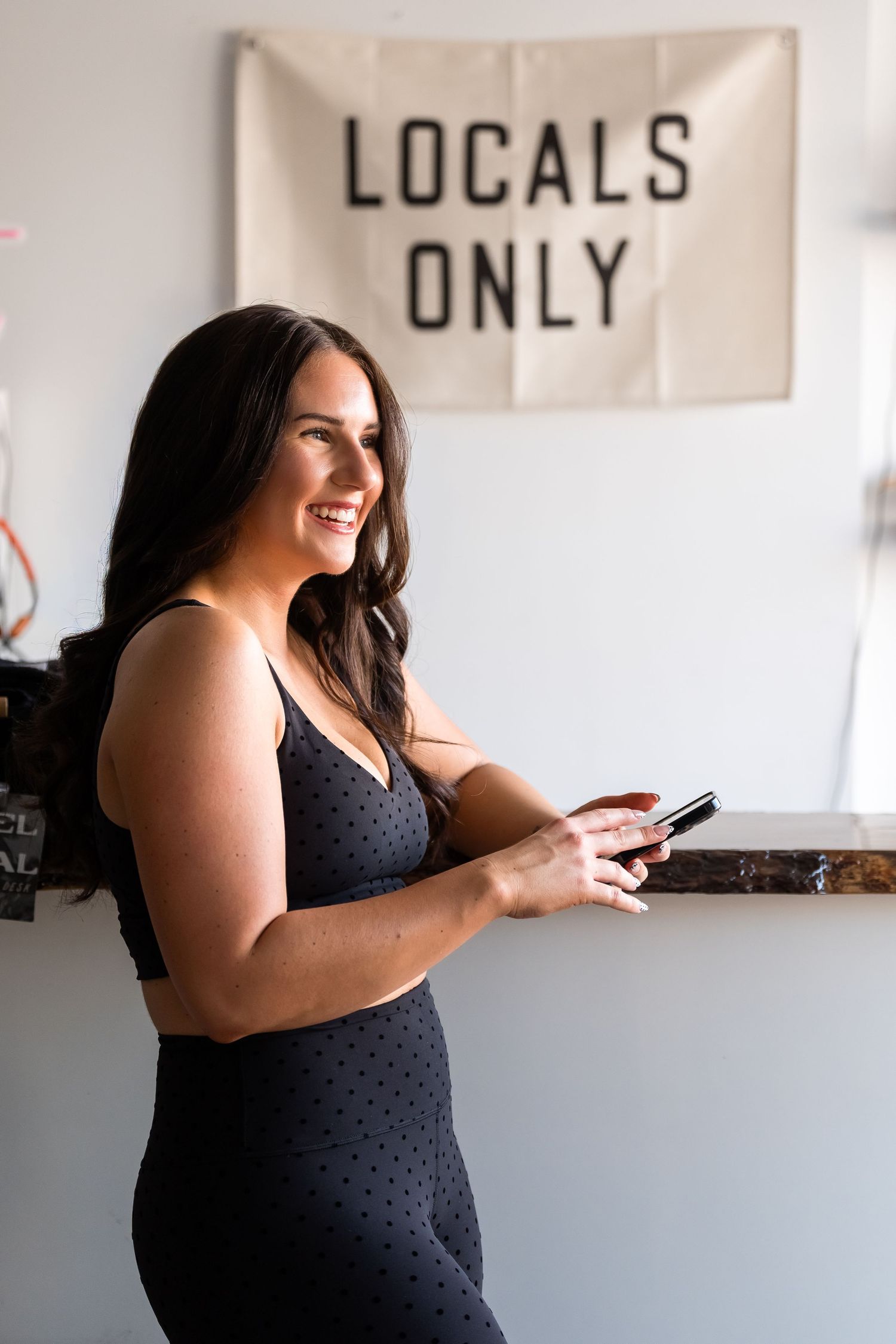 Woman in black polka dot dress smiles while using phone in bright interior space with wall signage.