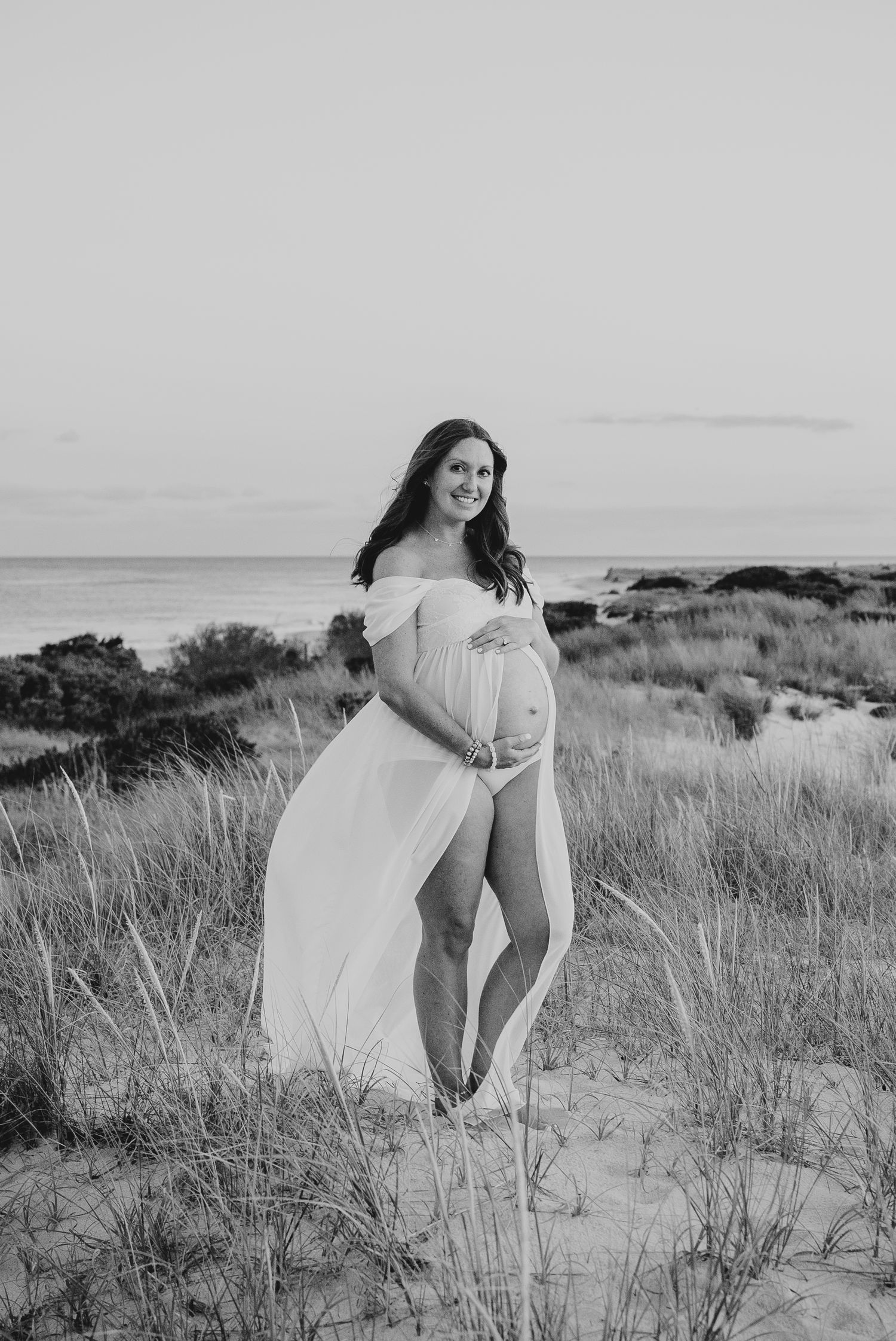 Artistic maternity photo on a beach with flowing white dress and natural coastal scenery.