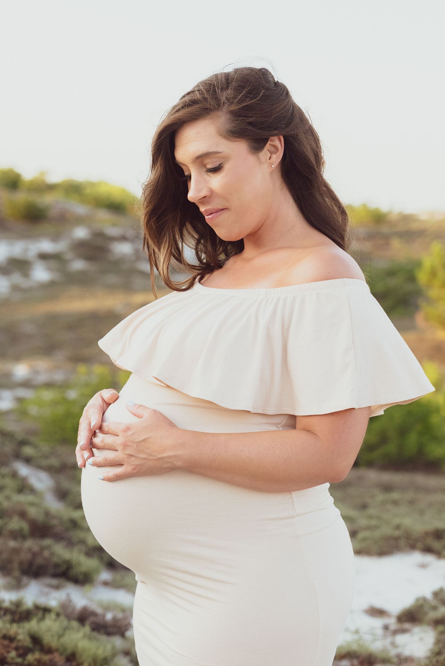 Woman in off-shoulder white dress cradling baby bump outdoors.