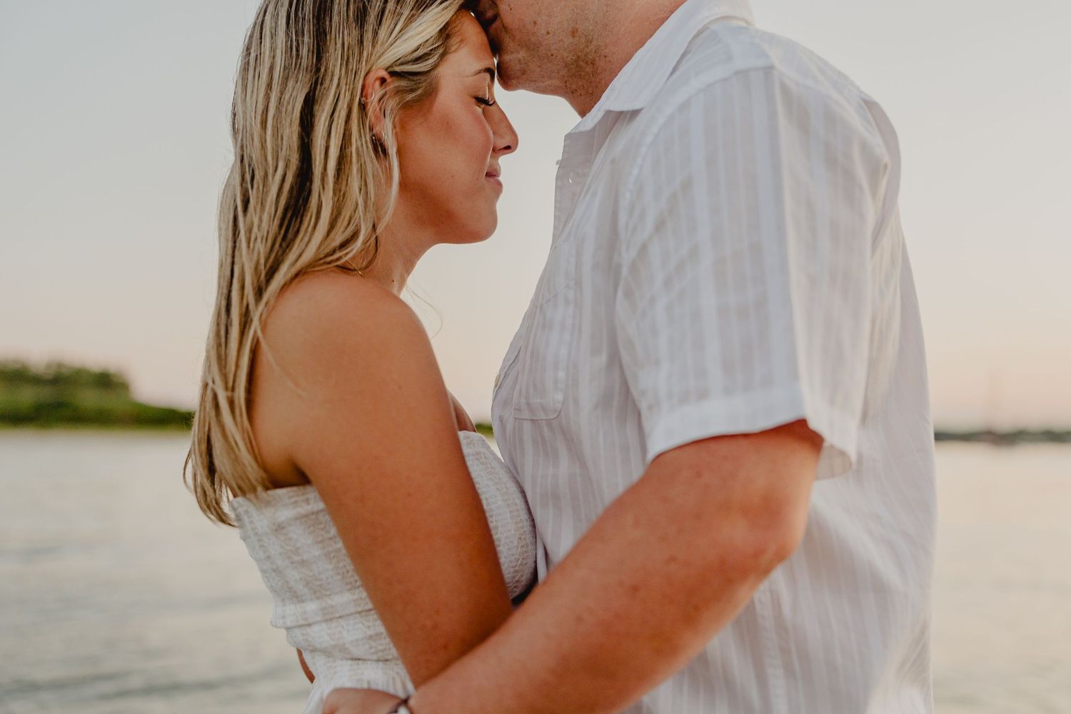 Couple sharing a romantic moment near the water at sunset, woman in white dress embracing man in striped shirt.